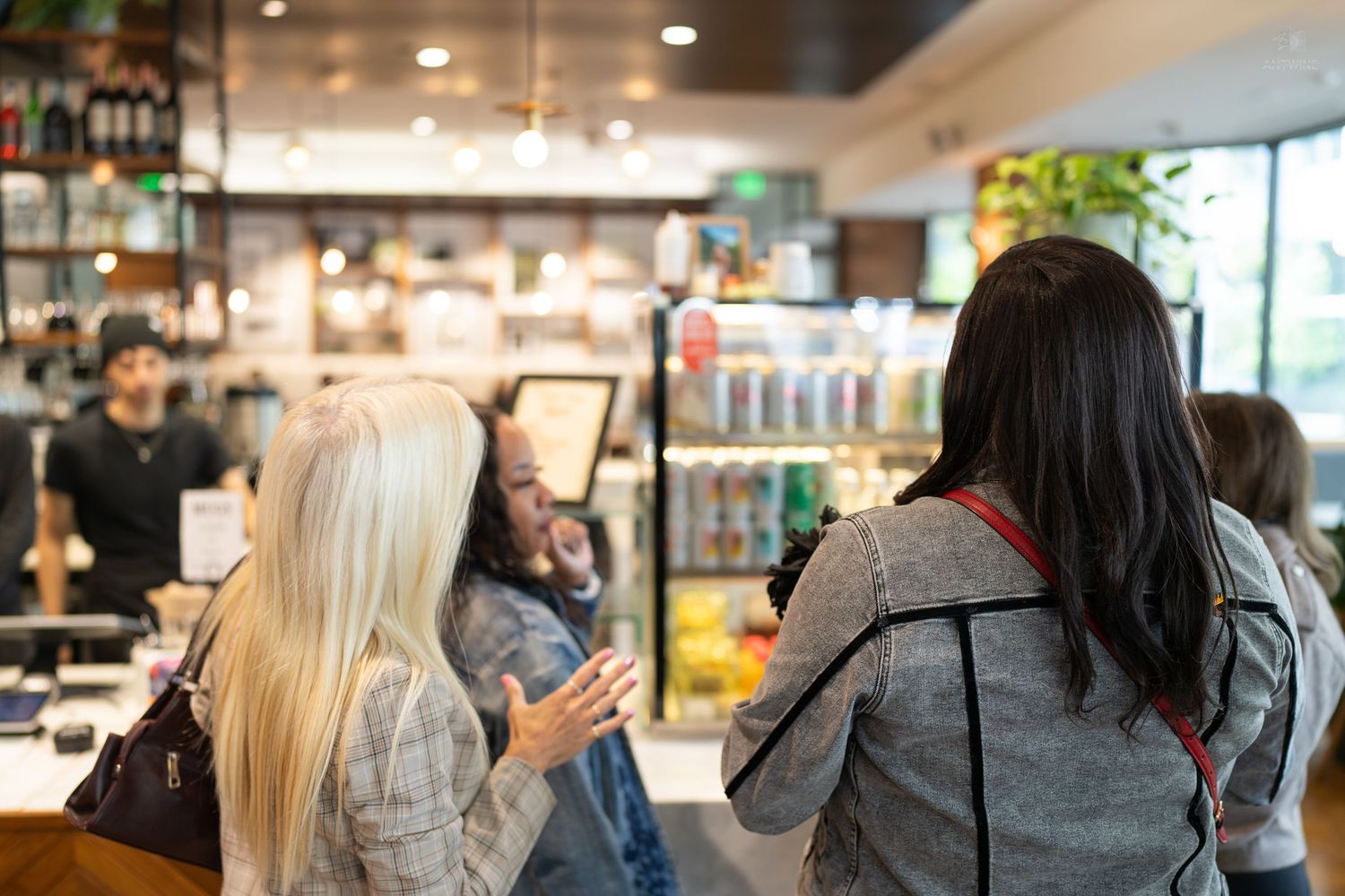 Women chatting at a modern cafe counter with refrigerated display and menu boards in background.