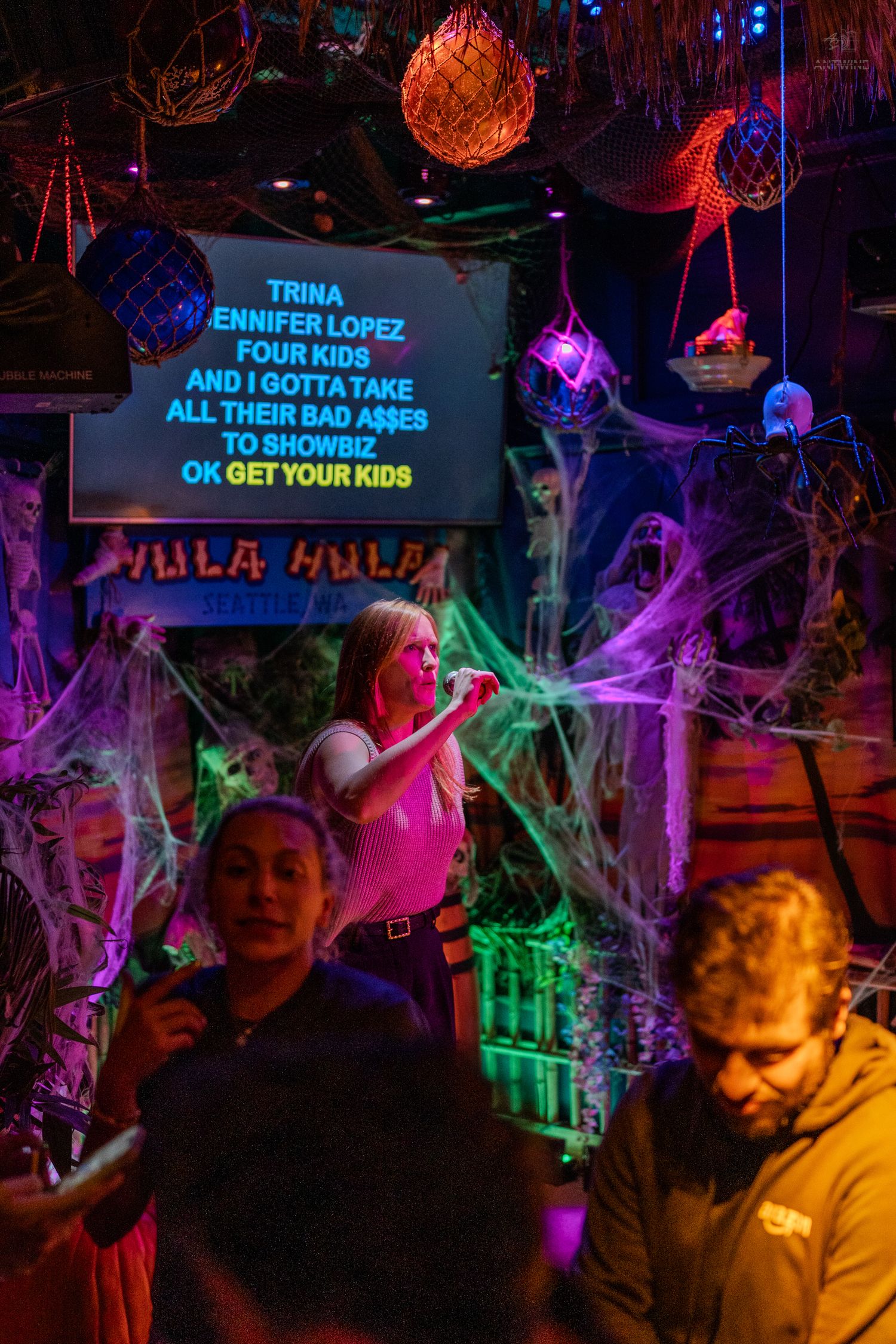 Blonde woman performing karaoke at Hula Hula bar surrounded by colorful neon lights and Halloween decor.