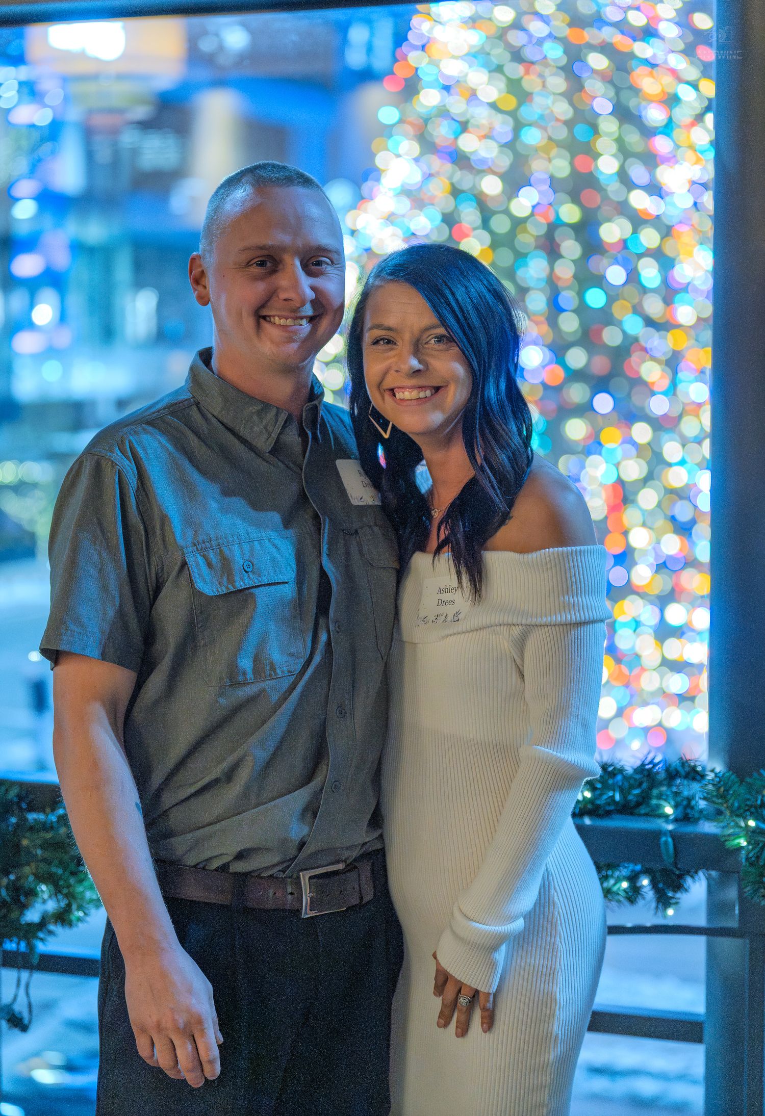 Smiling couple posing together in front of a colorful Christmas tree with bokeh lights backdrop.