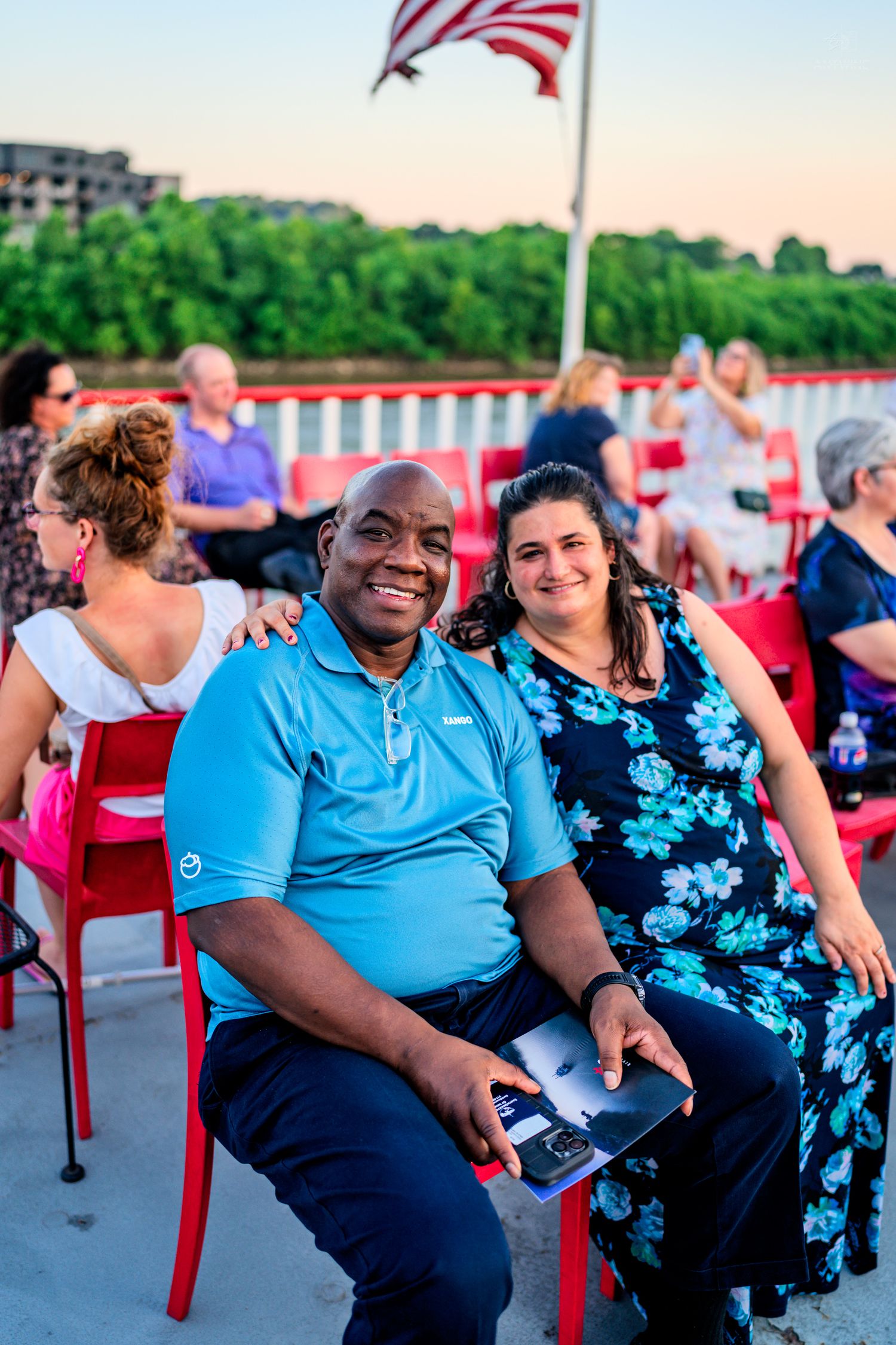 Smiling couple posing together in red chairs on an outdoor deck with an American flag at sunset.