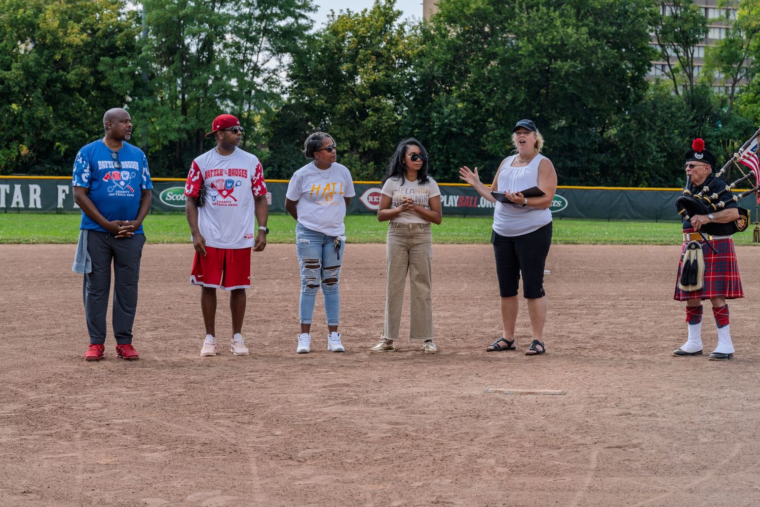 Six people gathered on a baseball field infield during a ceremonial event with a bagpiper present.