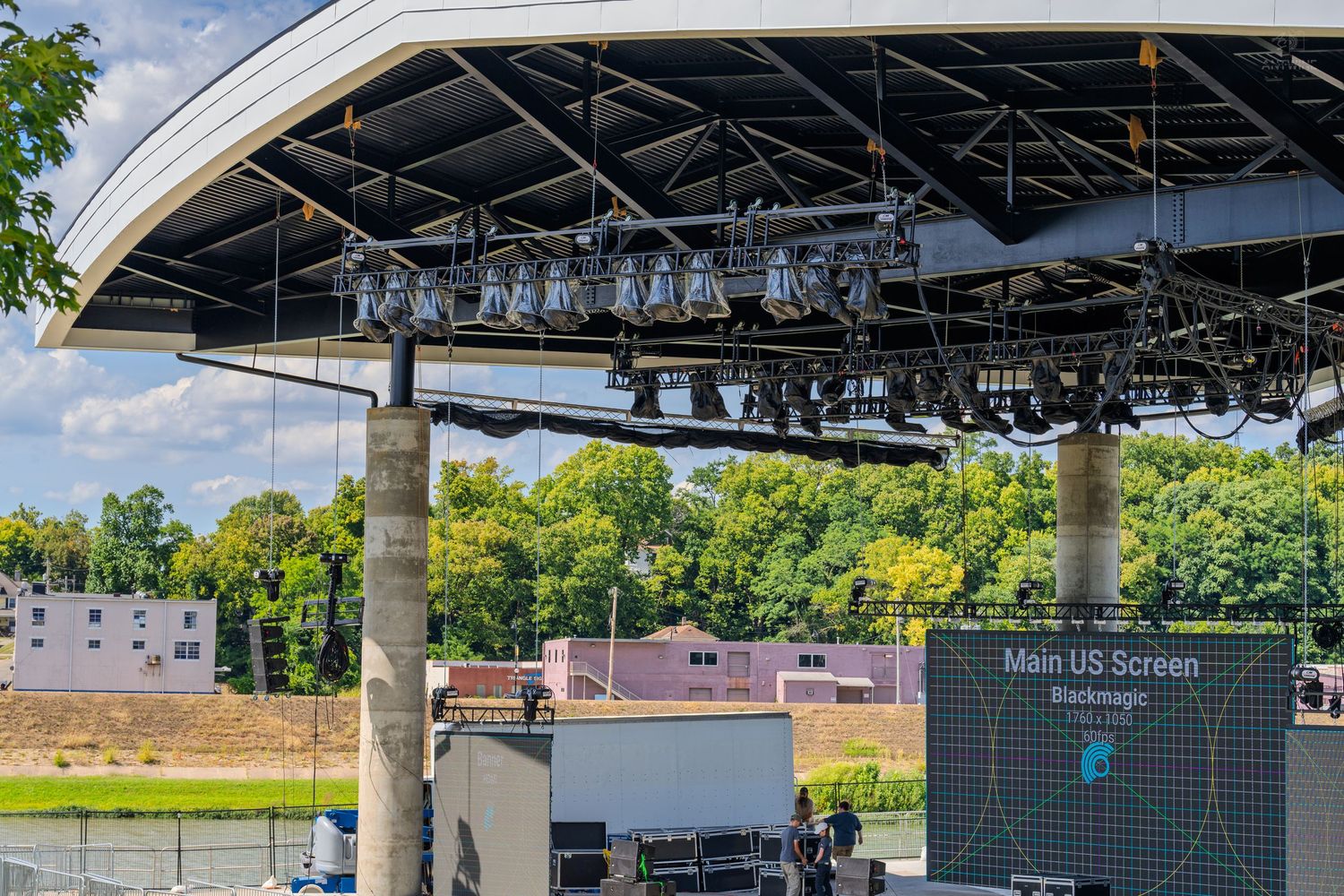 Outdoor concert stage with professional lighting rig and large LED screen under a curved roof canopy.