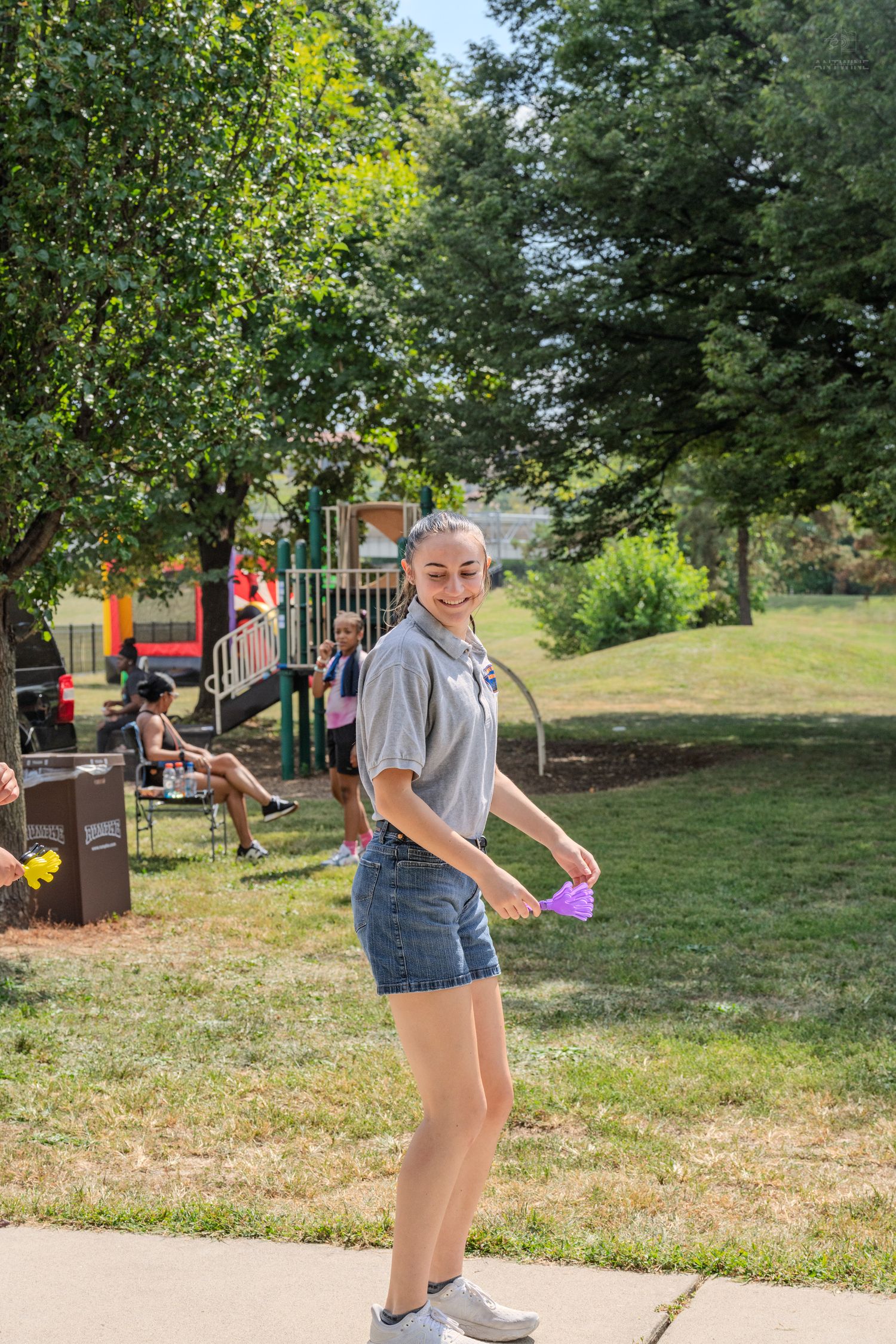 Smiling young woman in gray top and denim shorts holds purple cup in a sunny park with playground.