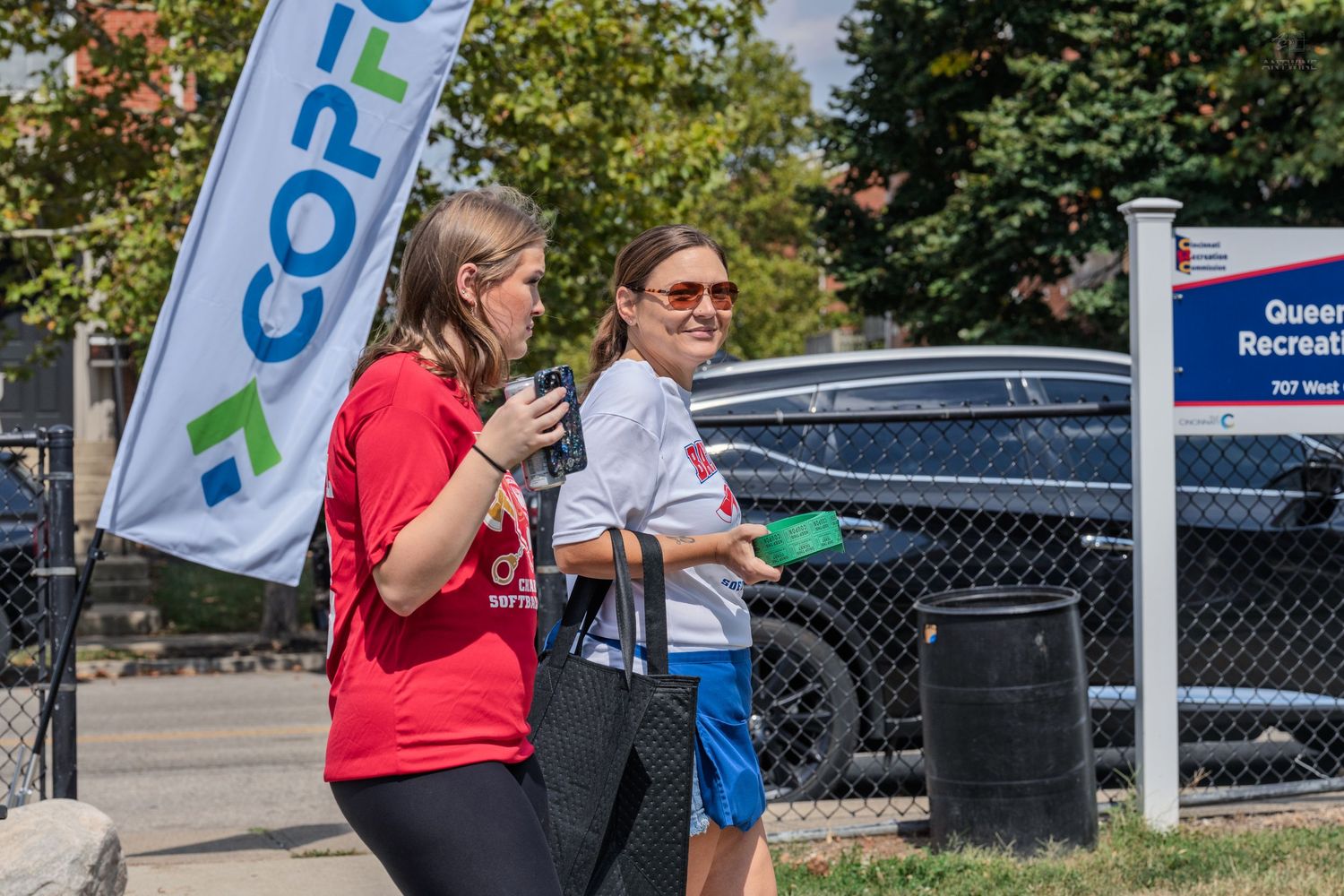 Two women in red and white shirts walk outdoors near a CopFirst banner and Queens Recreation sign on a sunny day.