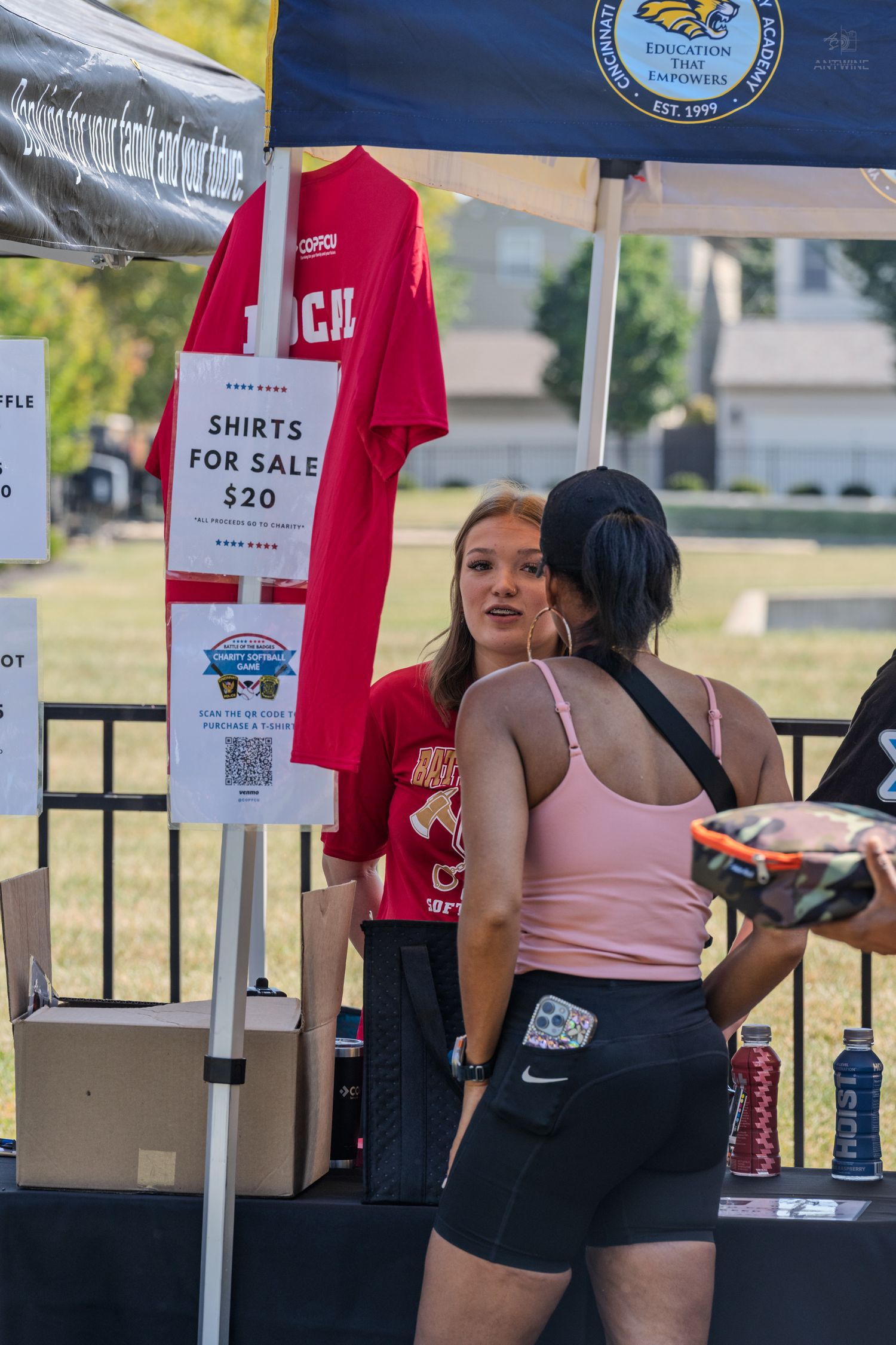 Young woman selling red shirts for $20 at an outdoor community event booth under a canopy.