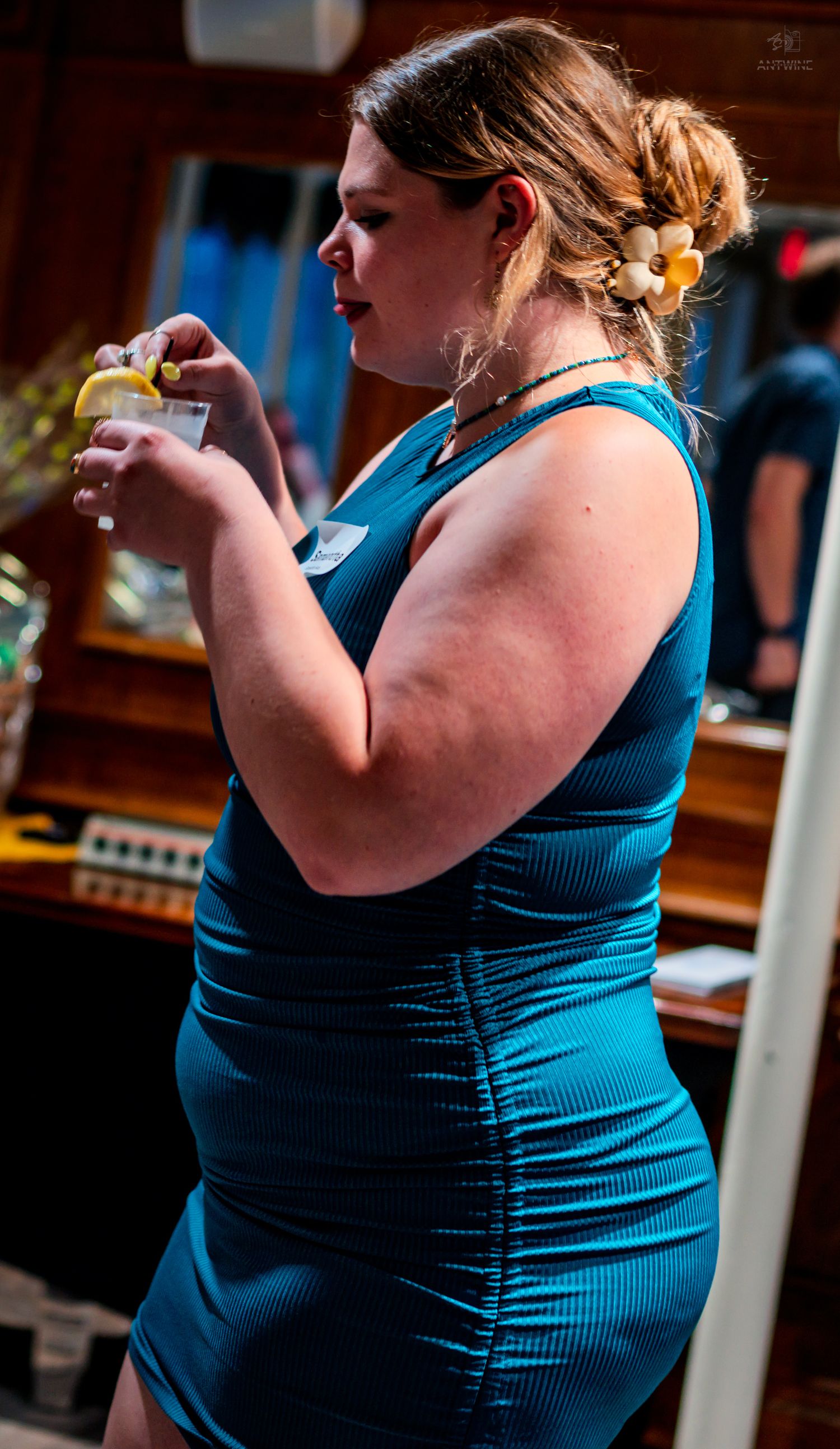A woman in a teal dress holds a cocktail glass with a lemon slice at an indoor event.
