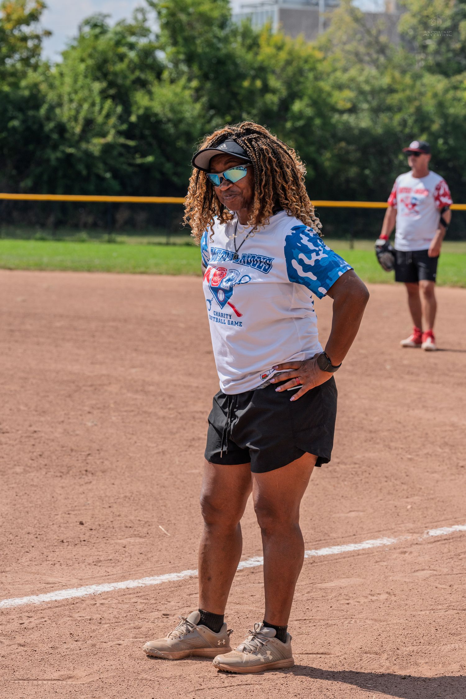 Woman in team jersey and visor standing confidently on softball field during charity game.