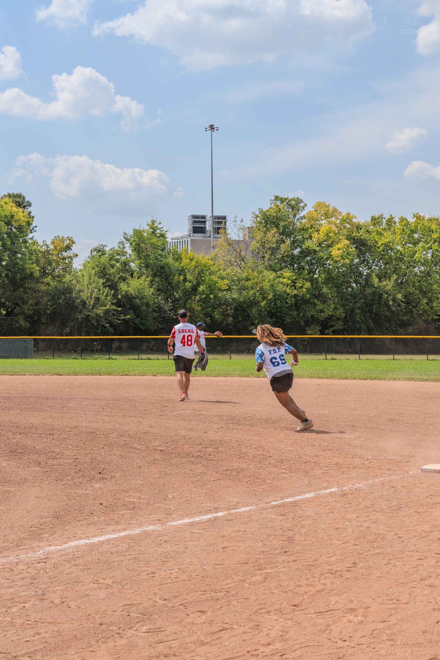 Two softball players run bases on dirt field during outdoor charity softball game under blue sky.
