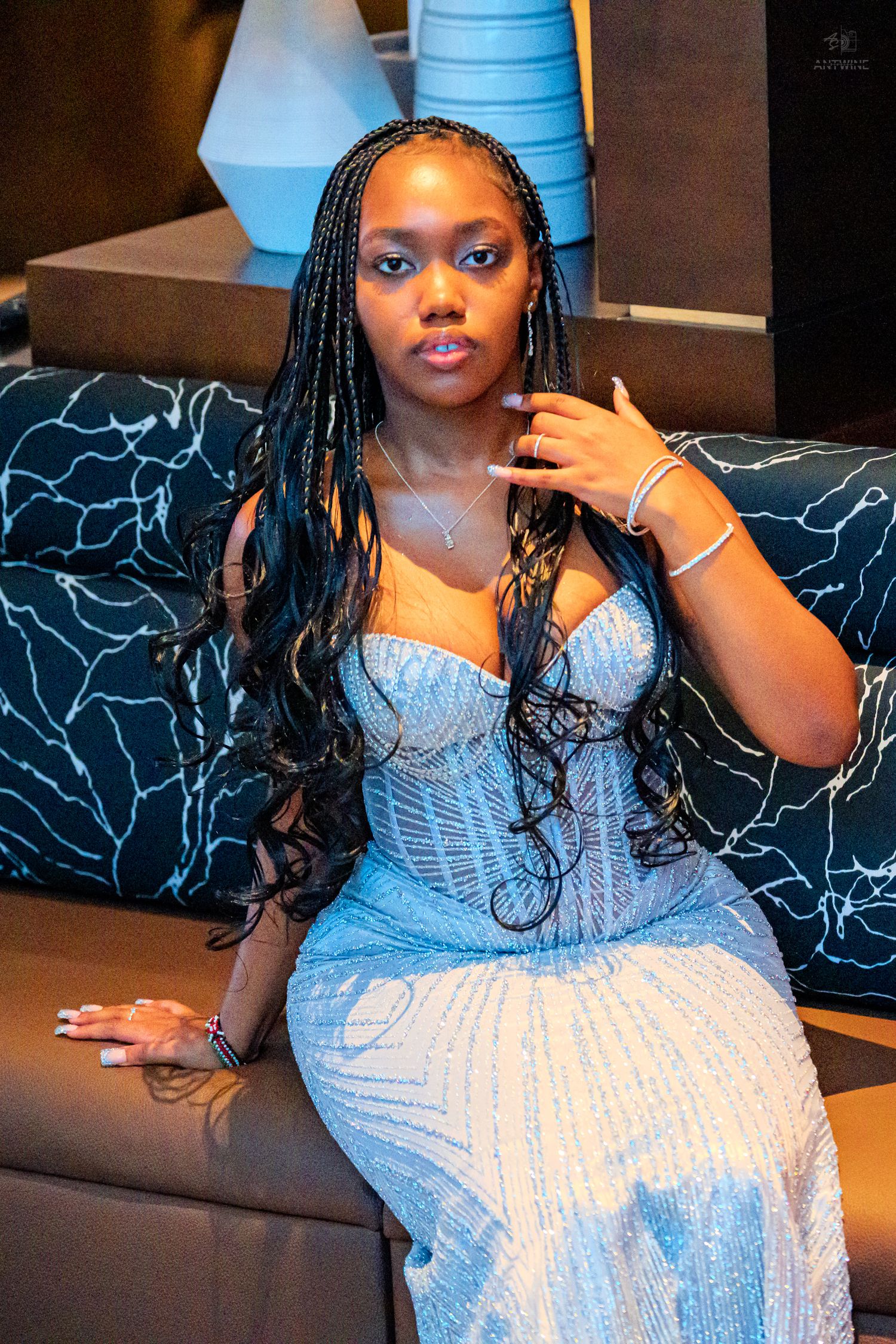 Young woman in sparkly blue corset gown with braids poses elegantly on dark sofa.