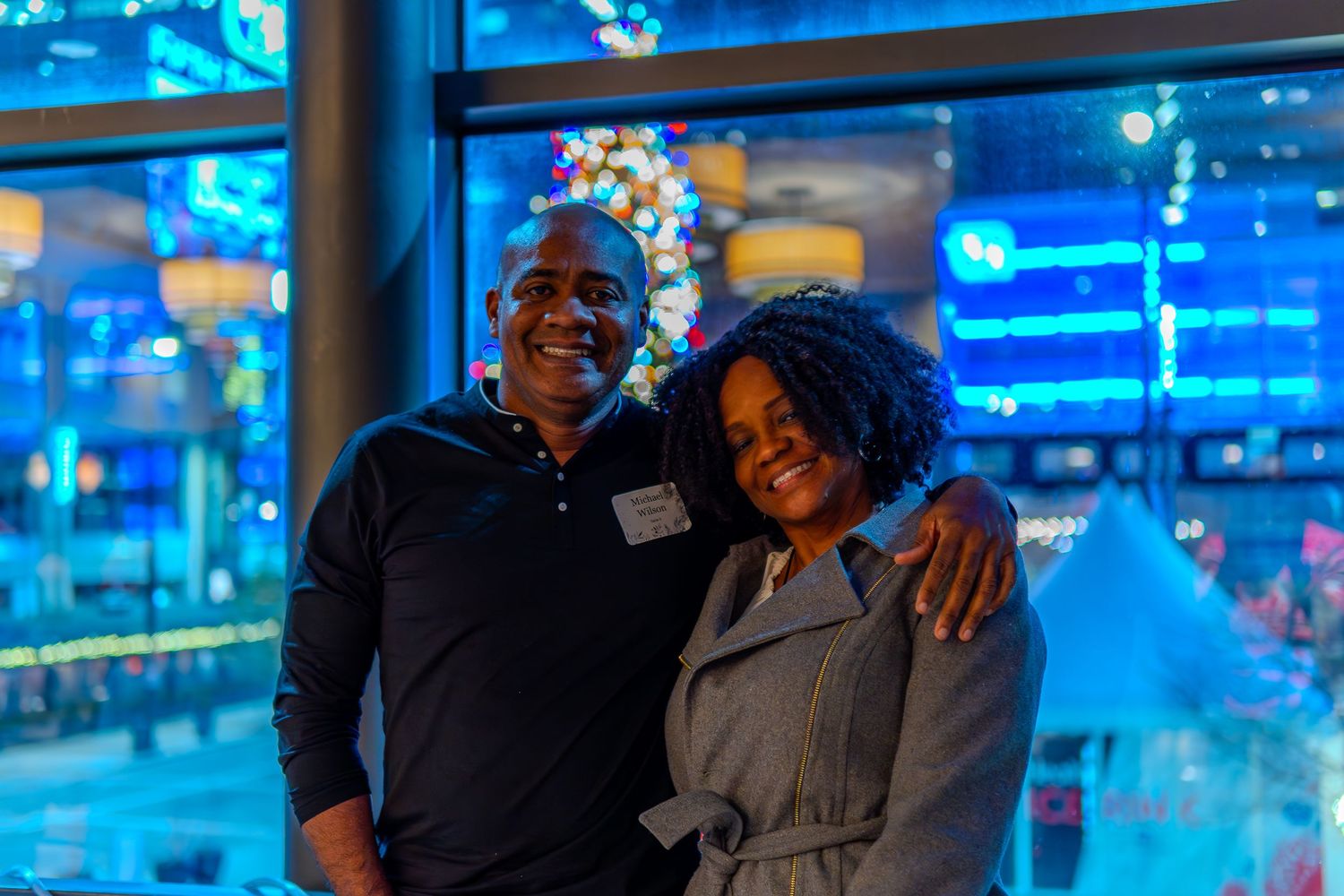 A smiling couple poses together at a festive holiday event with blue lights and a Christmas tree in the background.