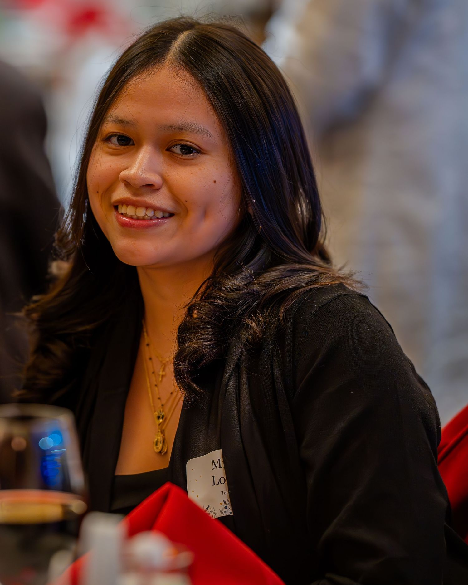 Young woman smiling warmly at a formal event, wearing a black blazer and gold necklace with a name tag.