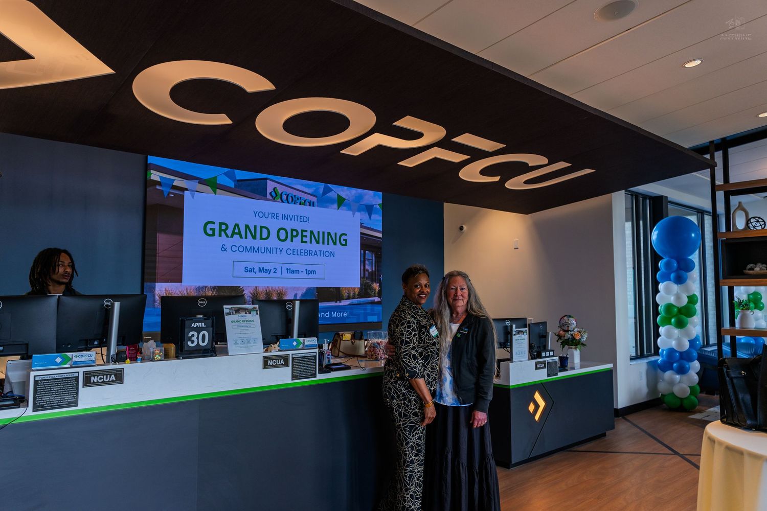 Two women stand at the counter of a COPCU credit union grand opening event with blue balloons in the background.
