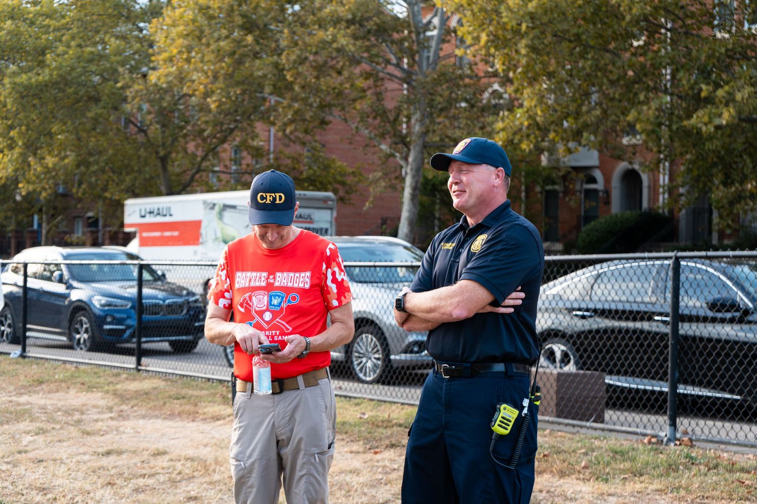 Two men, one in a CFD cap and red jersey, stand outdoors near a chain-link fence with parked cars in the background.