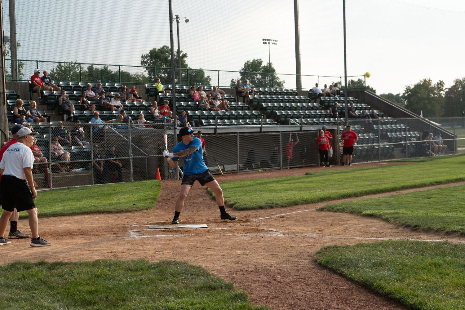 Batter swings at a softball game with spectators in bleachers under evening sky at an outdoor baseball diamond.