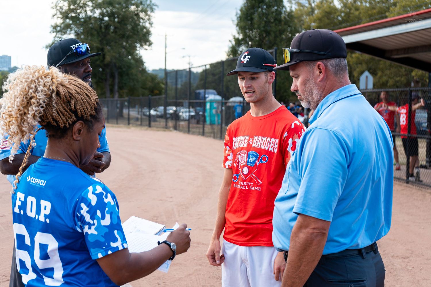 People converse at a youth baseball event on a sunny day at an outdoor sports field.