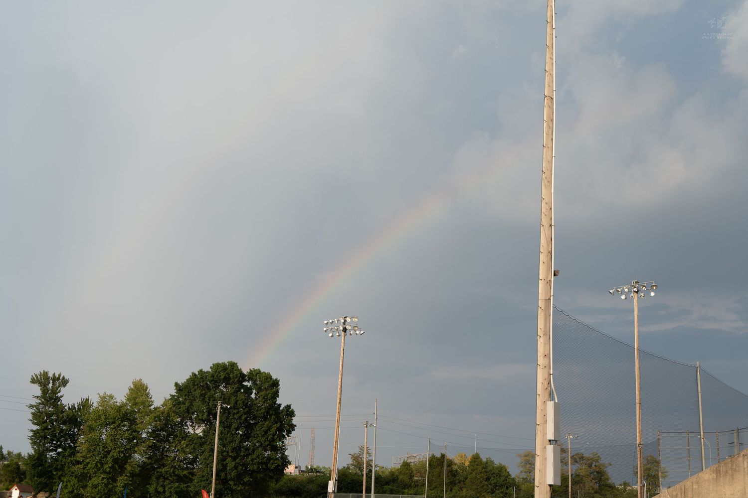 A vibrant rainbow arches over a sports field with tall light poles and lush green trees under a cloudy sky.