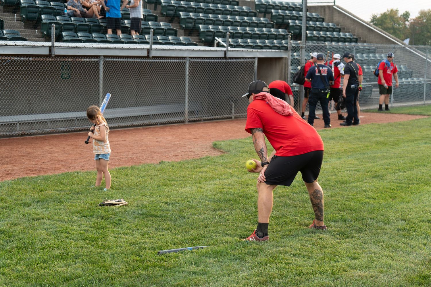 A child bats while an adult pitches during a casual baseball game on a green field near stadium bleachers.