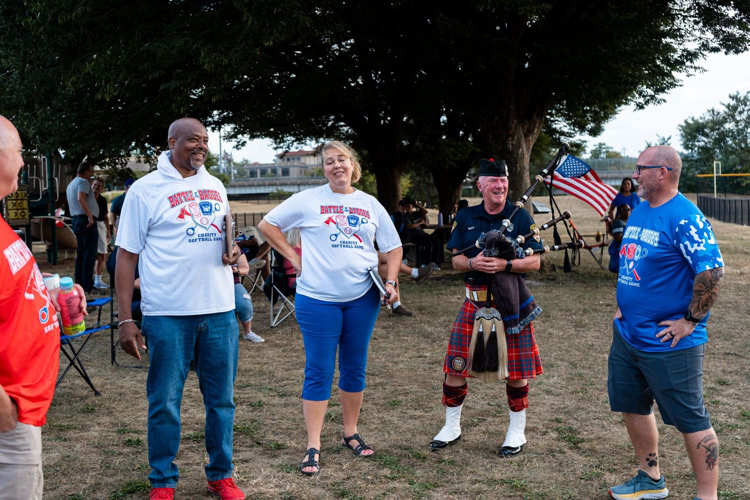 Group of people in patriotic shirts and a bagpiper in kilts gathered outdoors at a community event.