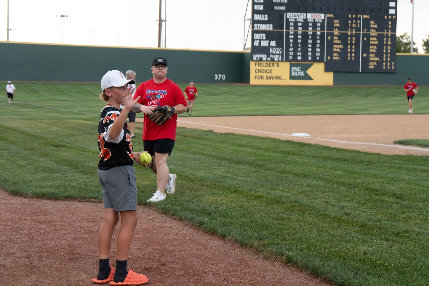 Two players practice throwing on baseball field with scoreboard visible in background during evening game.