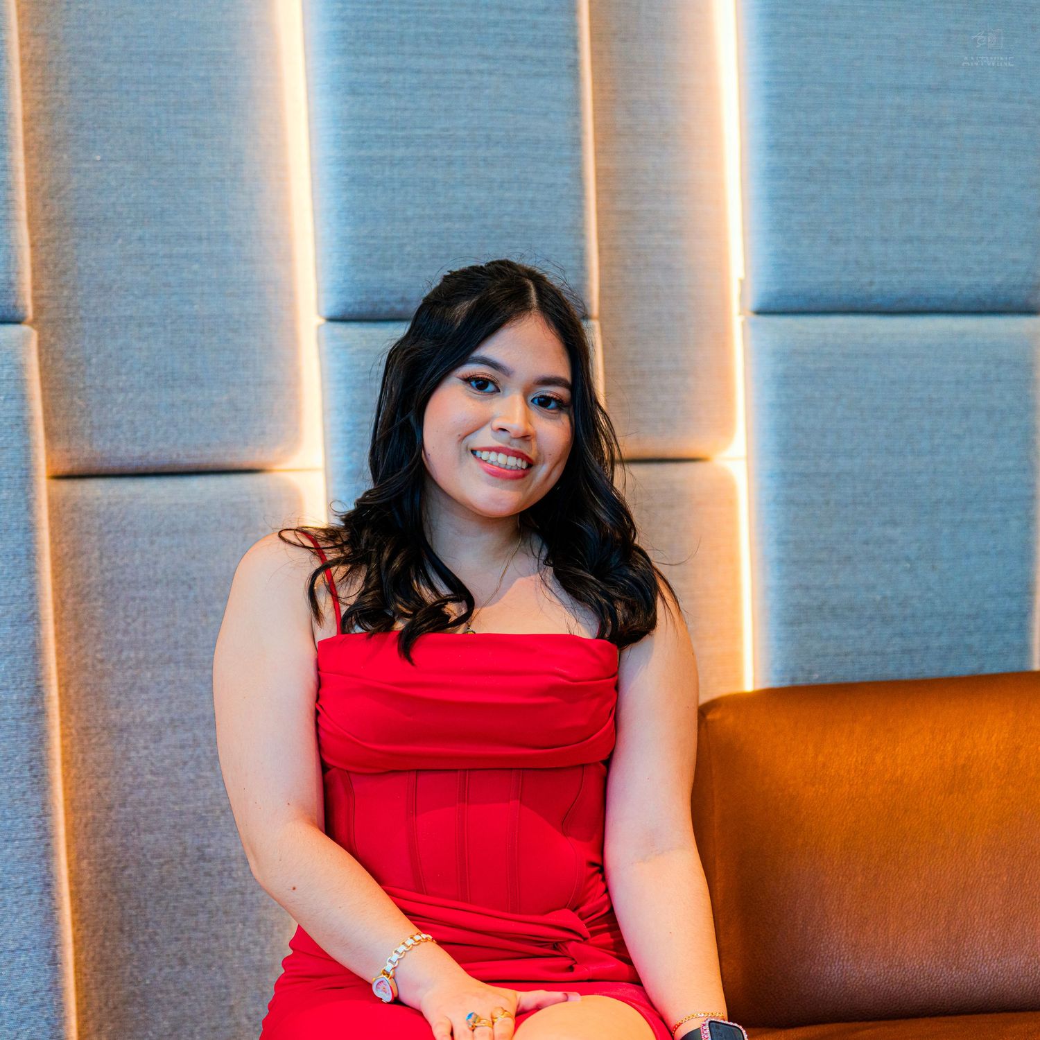 Smiling young woman in red strapless dress seated on brown sofa in modern venue.