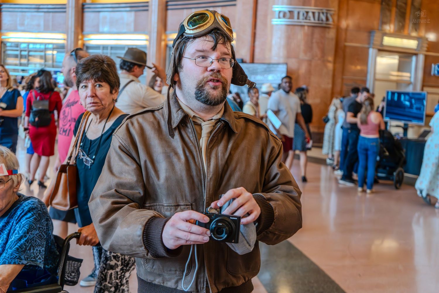 Man in vintage aviator jacket and goggles holds camera inside a busy train station hall.