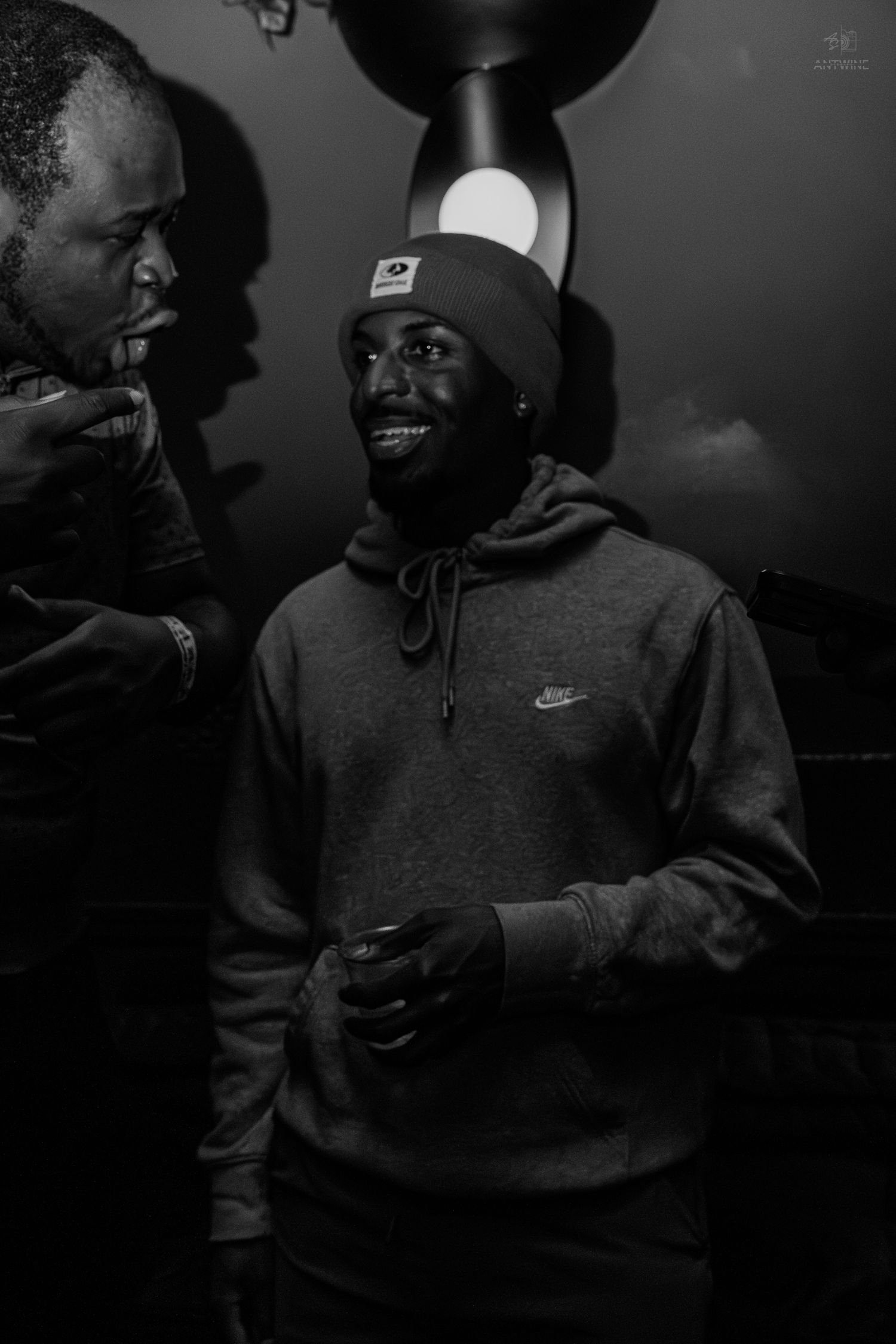 Smiling man wearing a Nike hoodie and beanie hat at a dimly lit social gathering.