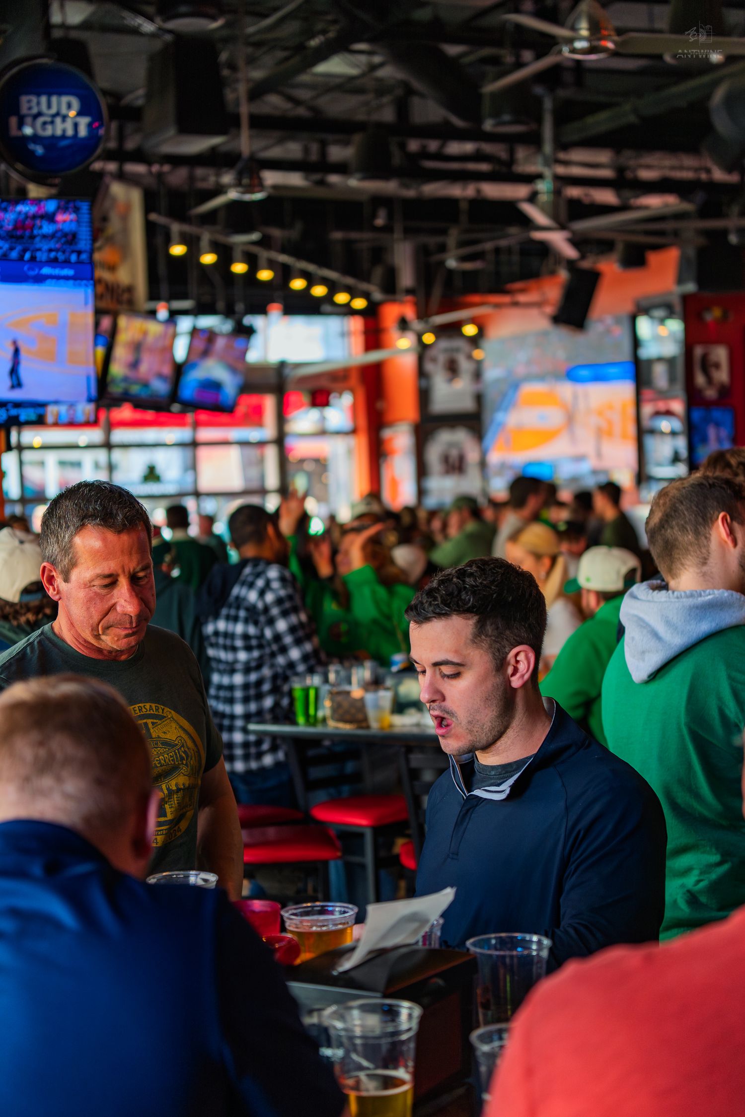 Crowded sports bar with Bud Light signage, multiple TVs, and patrons in green shirts enjoying drinks.