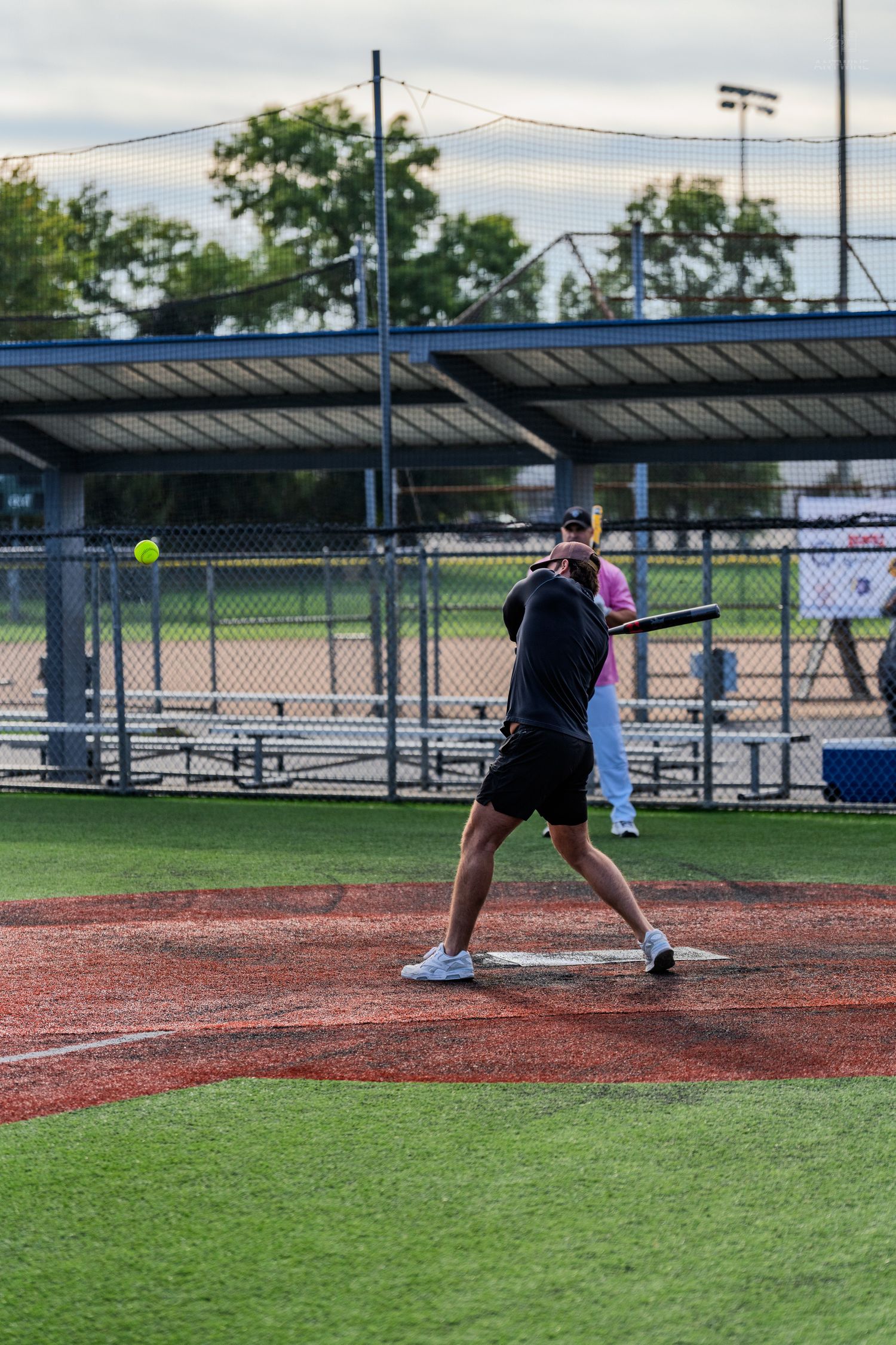 A batter swings at a yellow softball during a recreational league game at an outdoor baseball field.