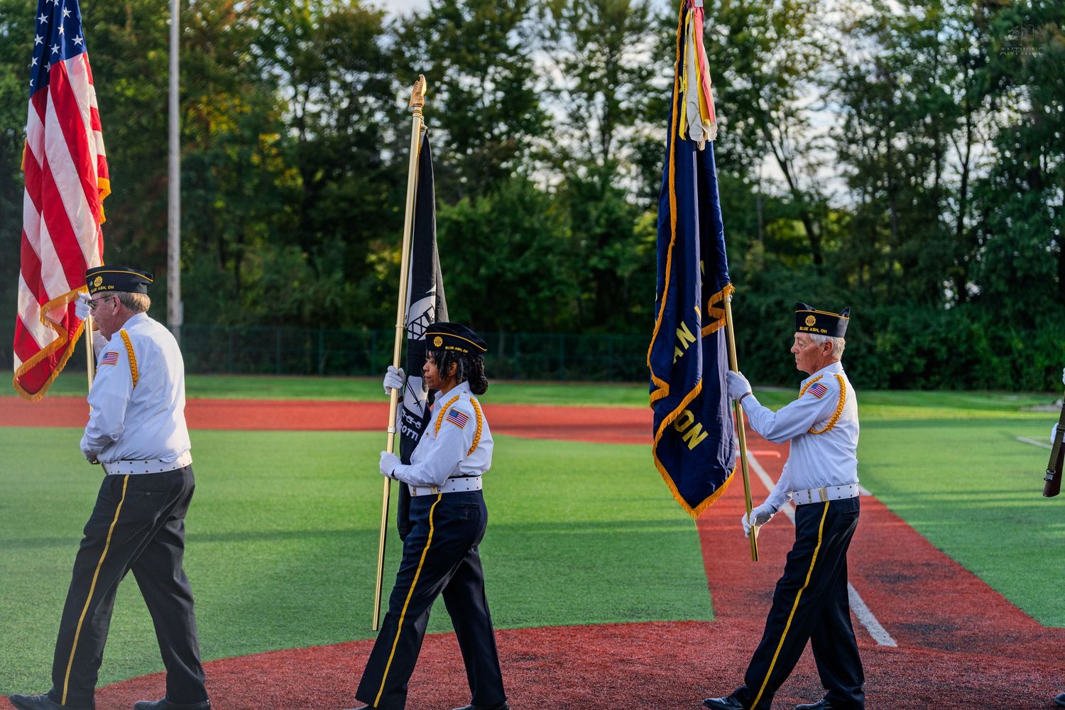 Military color guard carrying American and Navy flags on a red track at outdoor ceremony.