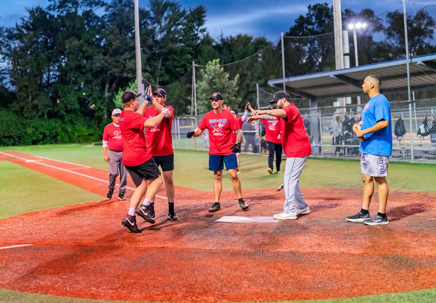 Softball players in red uniforms celebrate with high-fives on the field during a night game under bright lights.