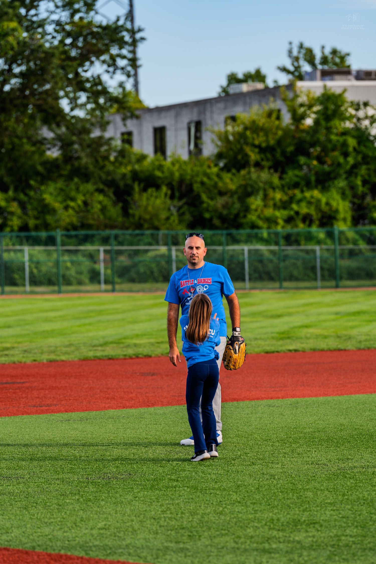 A man in a blue shirt coaches a young girl holding a baseball glove on a green softball field.