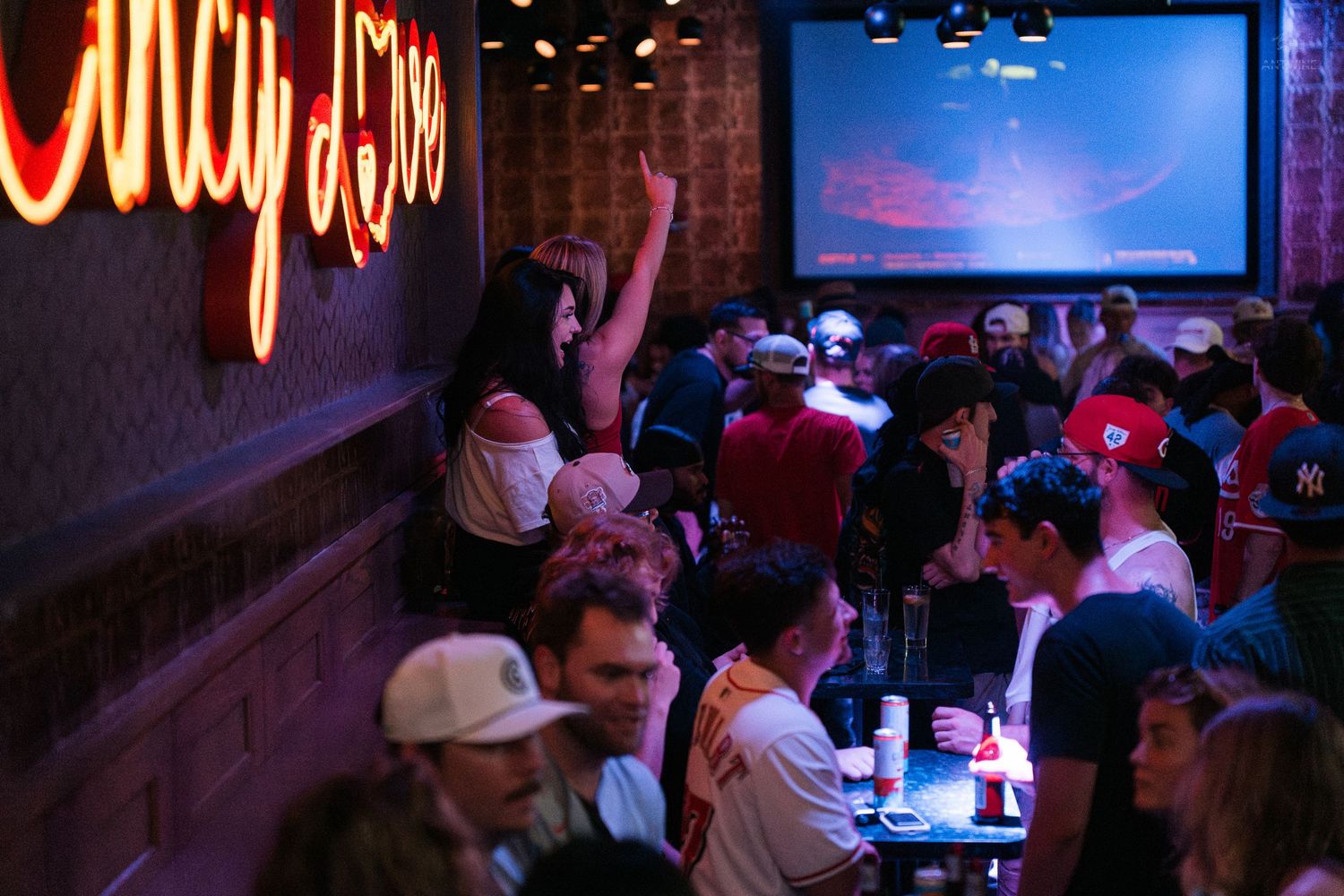 Crowded sports bar with neon signs, large projection screen, and lively patrons enjoying drinks and conversation.