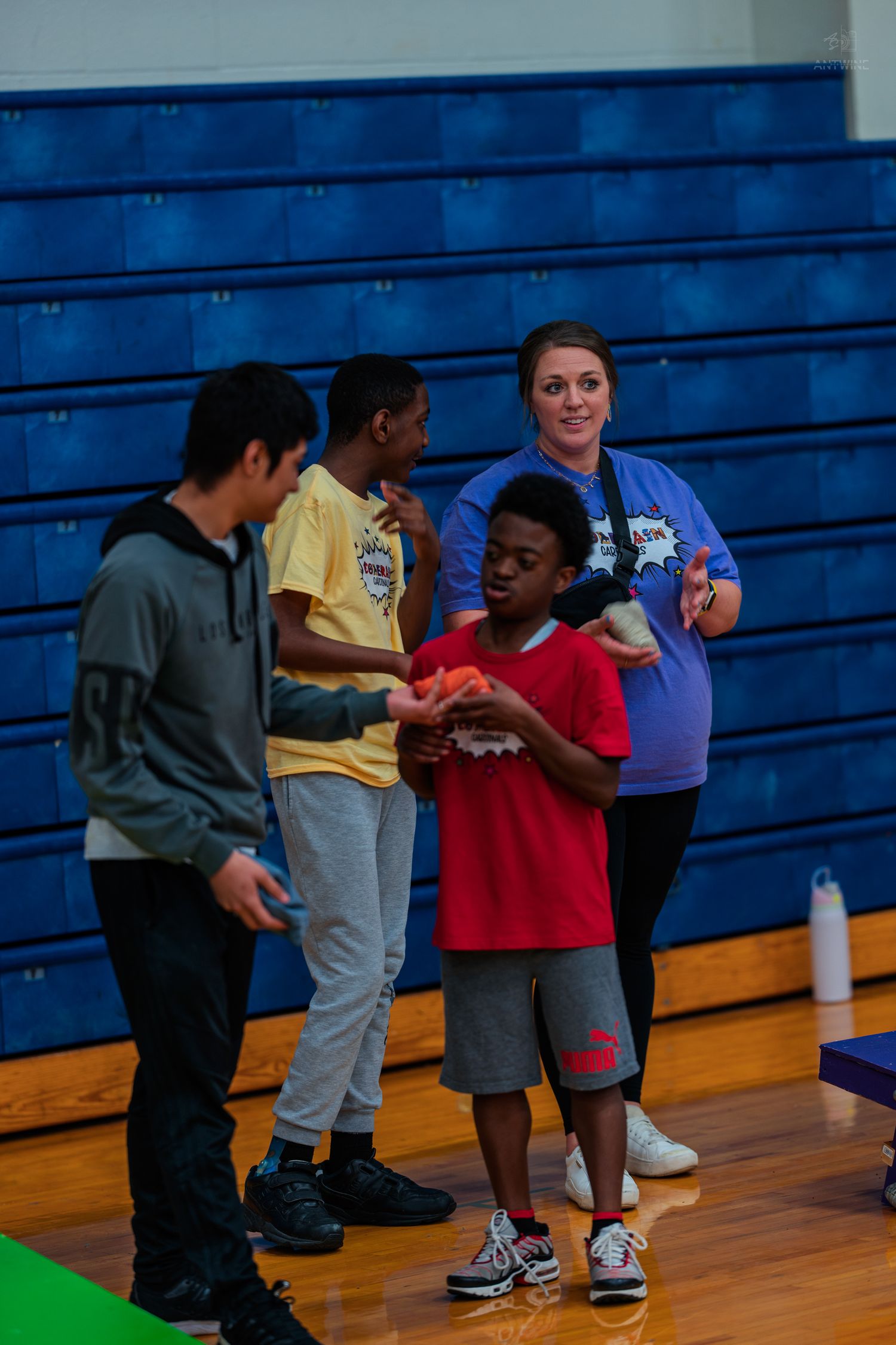 Students and a teacher participate in a gym activity inside a school gymnasium with blue bleachers.