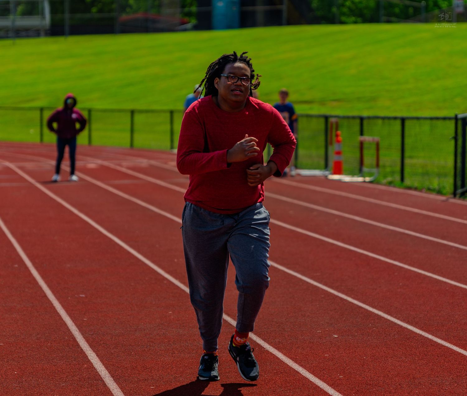 A person in a red shirt and gray pants runs on a red athletic track with green grass in the background.