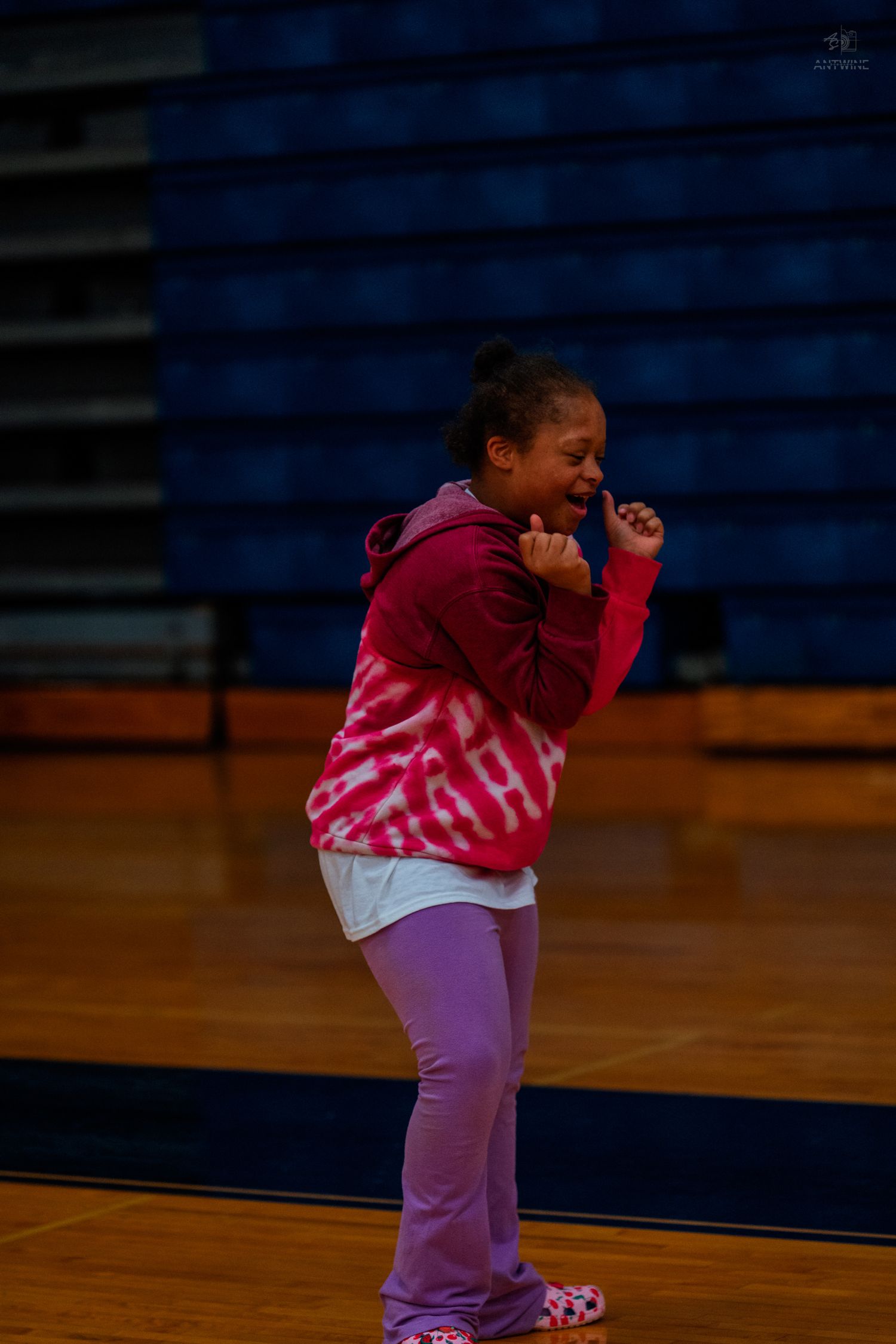 Young girl in pink tie-dye hoodie and lavender pants laughing joyfully in a gymnasium.