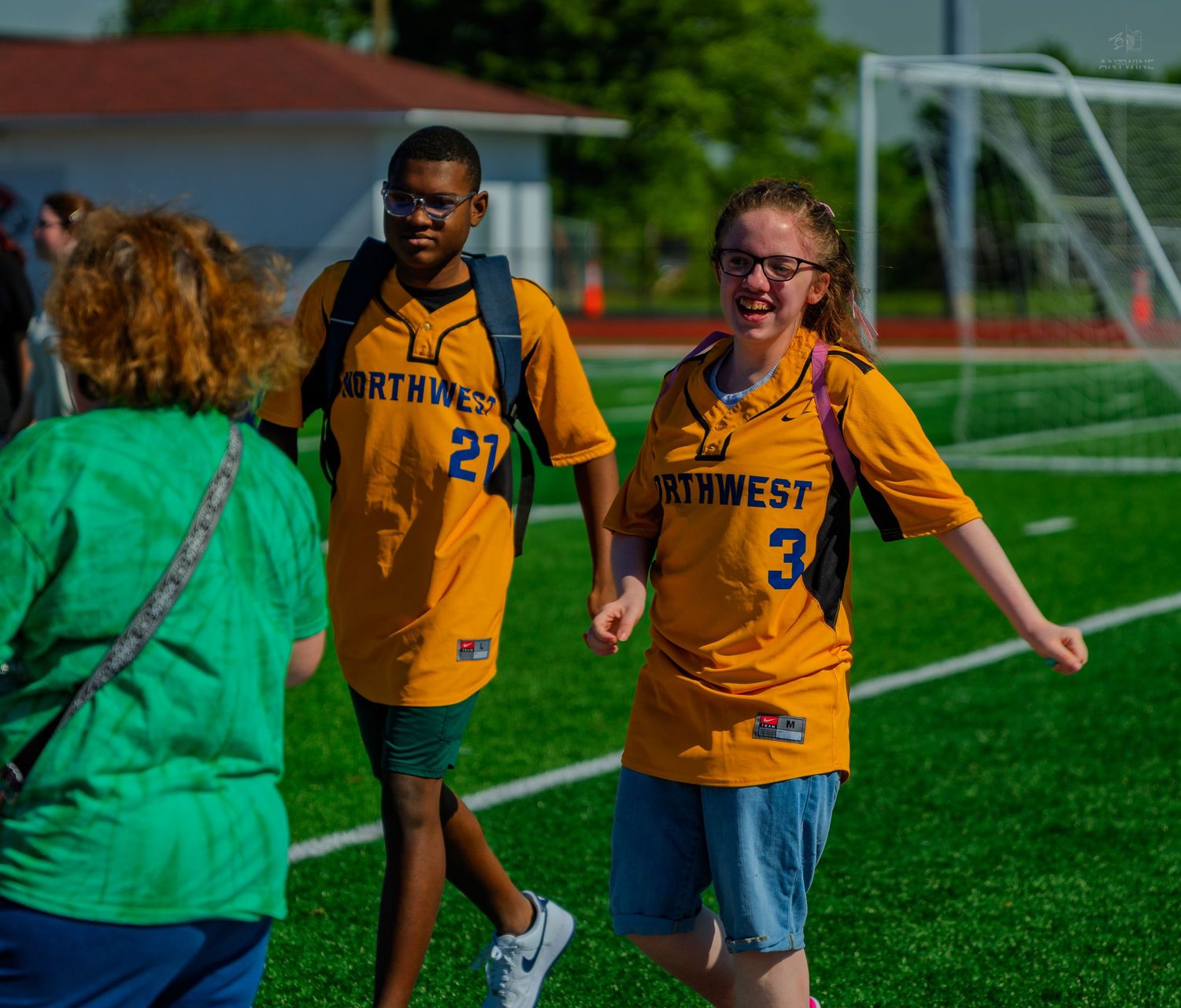 Two students in Northwest jerseys numbered 27 and 34 smile and interact on a green turf soccer field.