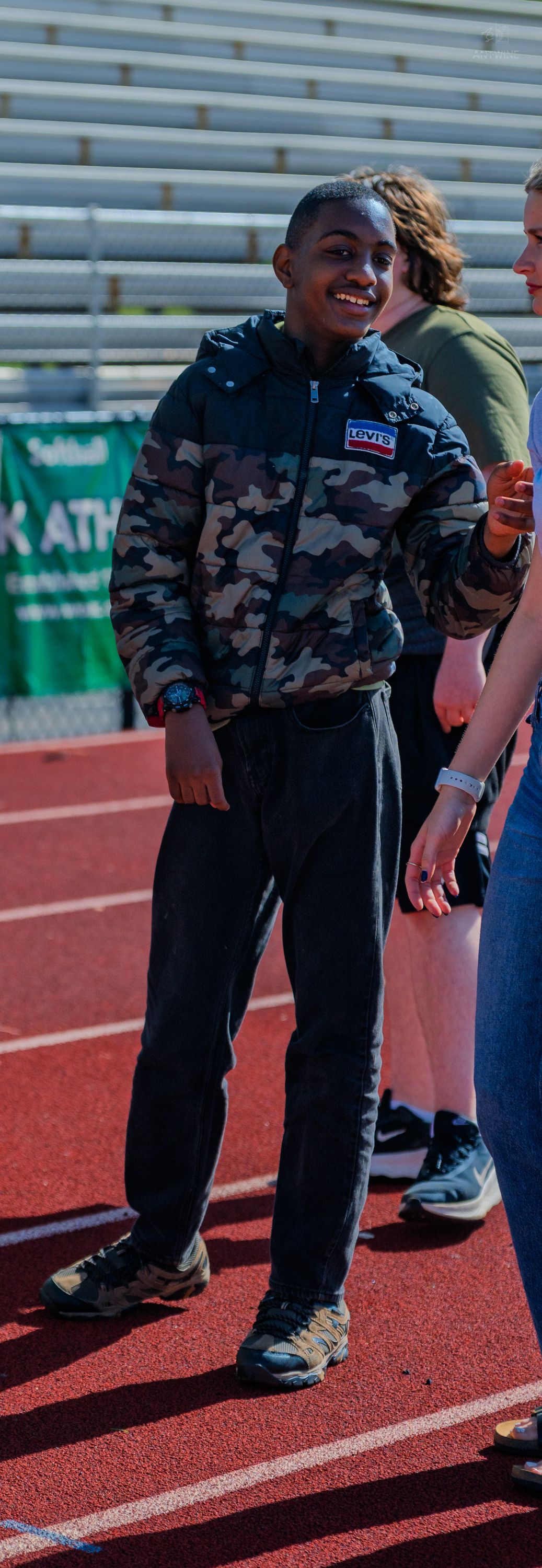 Smiling young boy wearing a camo puffer jacket and black pants standing on a red running track at a stadium.