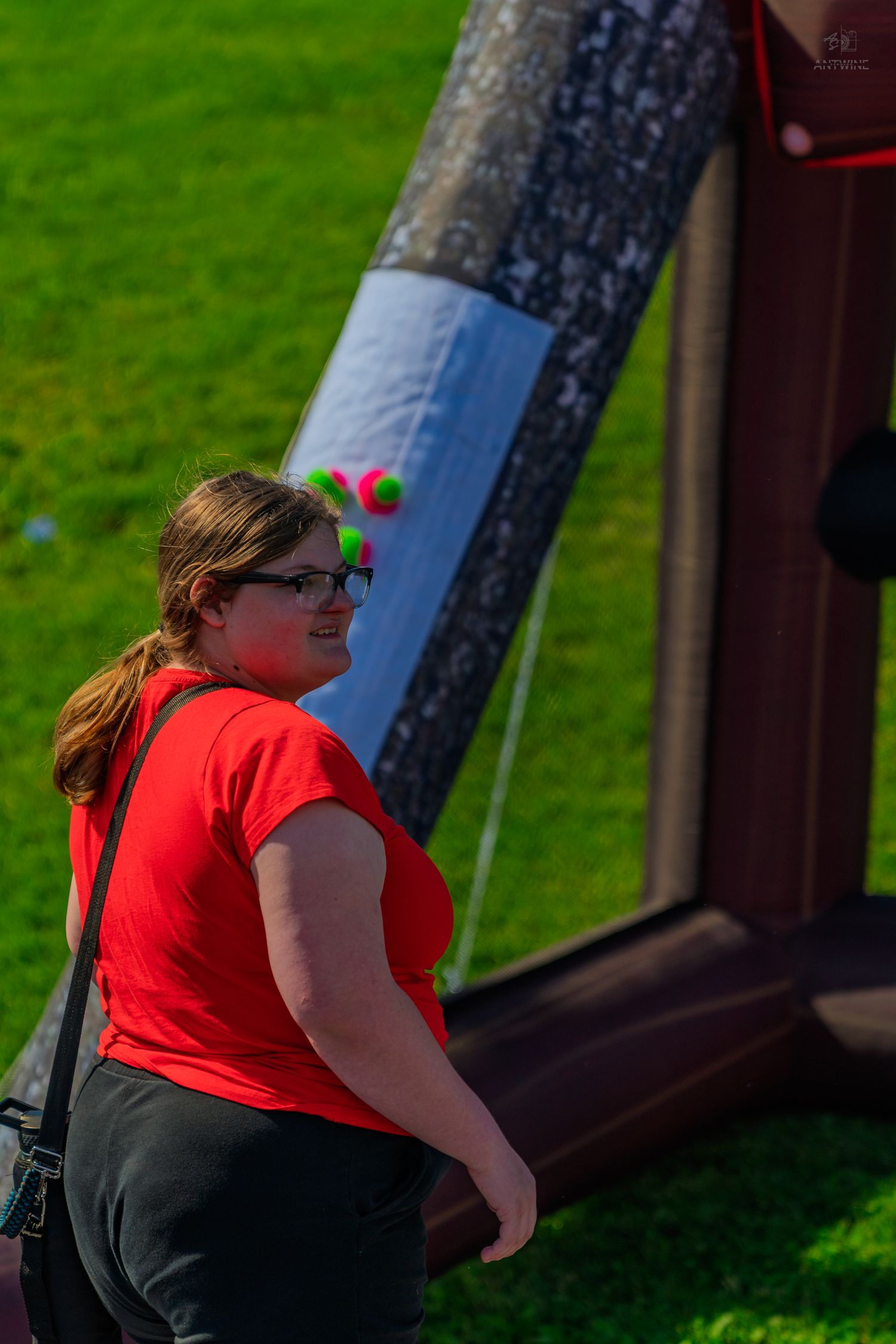 Woman in red shirt with glasses plays carnival game on green grass lawn at outdoor fair event.