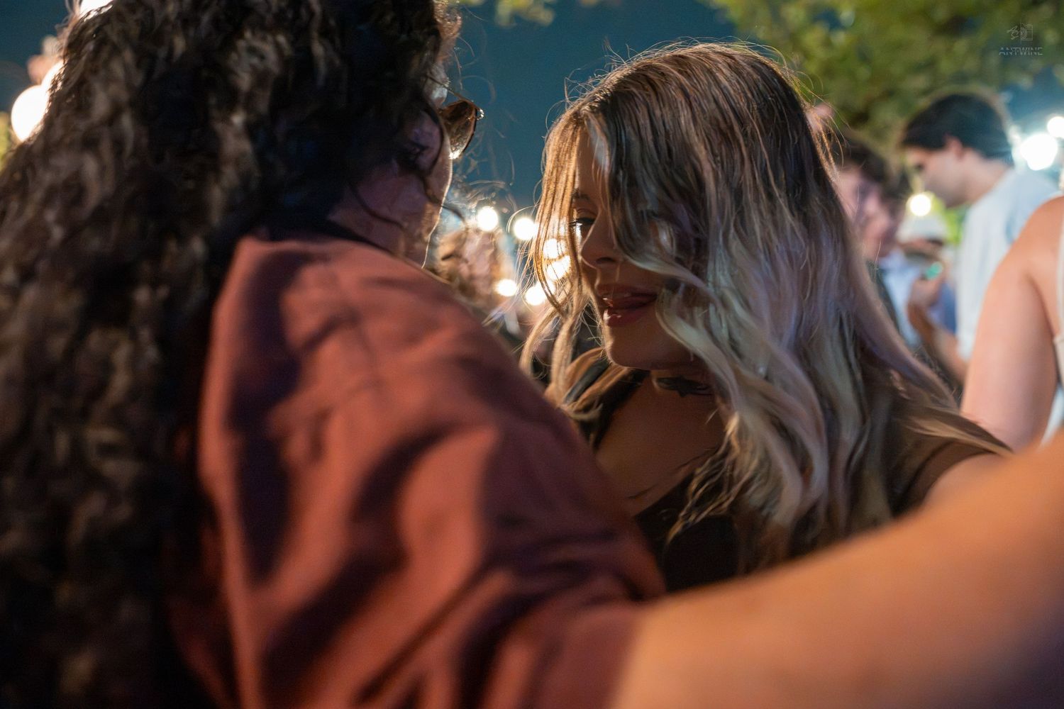 Two women embrace closely at an outdoor evening event, with soft bokeh lights in the background.