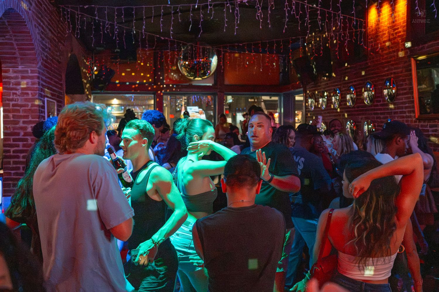 People dancing in a lively bar with colorful green and red lights, a disco ball, and brick walls.