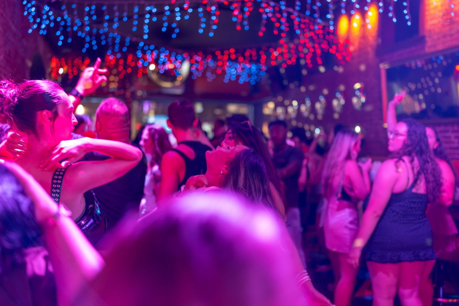 Crowd of people dancing in a vibrant nightclub with pink and blue neon lights and festive decorations.