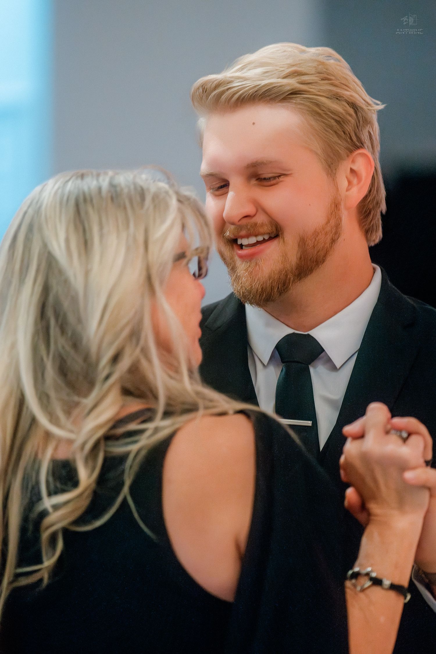 Smiling blond bearded man in black suit dances closely with blond woman in black dress at formal event.