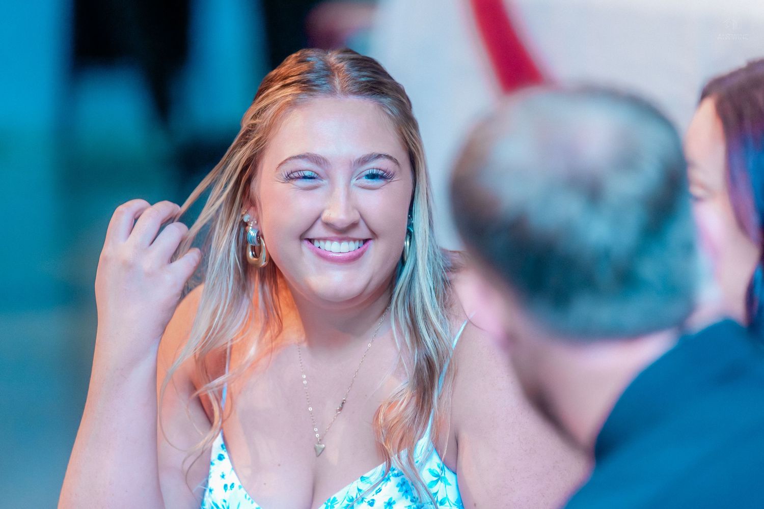 Smiling young blonde woman in floral dress touching her hair at a social event.
