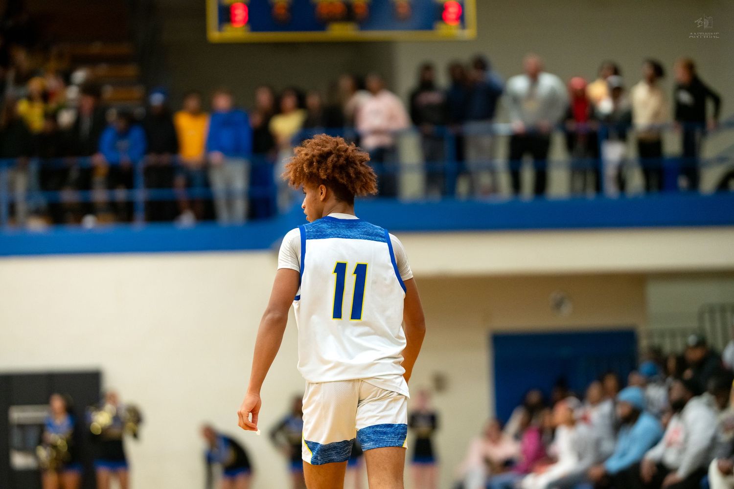 Basketball player #11 in blue and white uniform faces away toward crowded bleachers during indoor game.