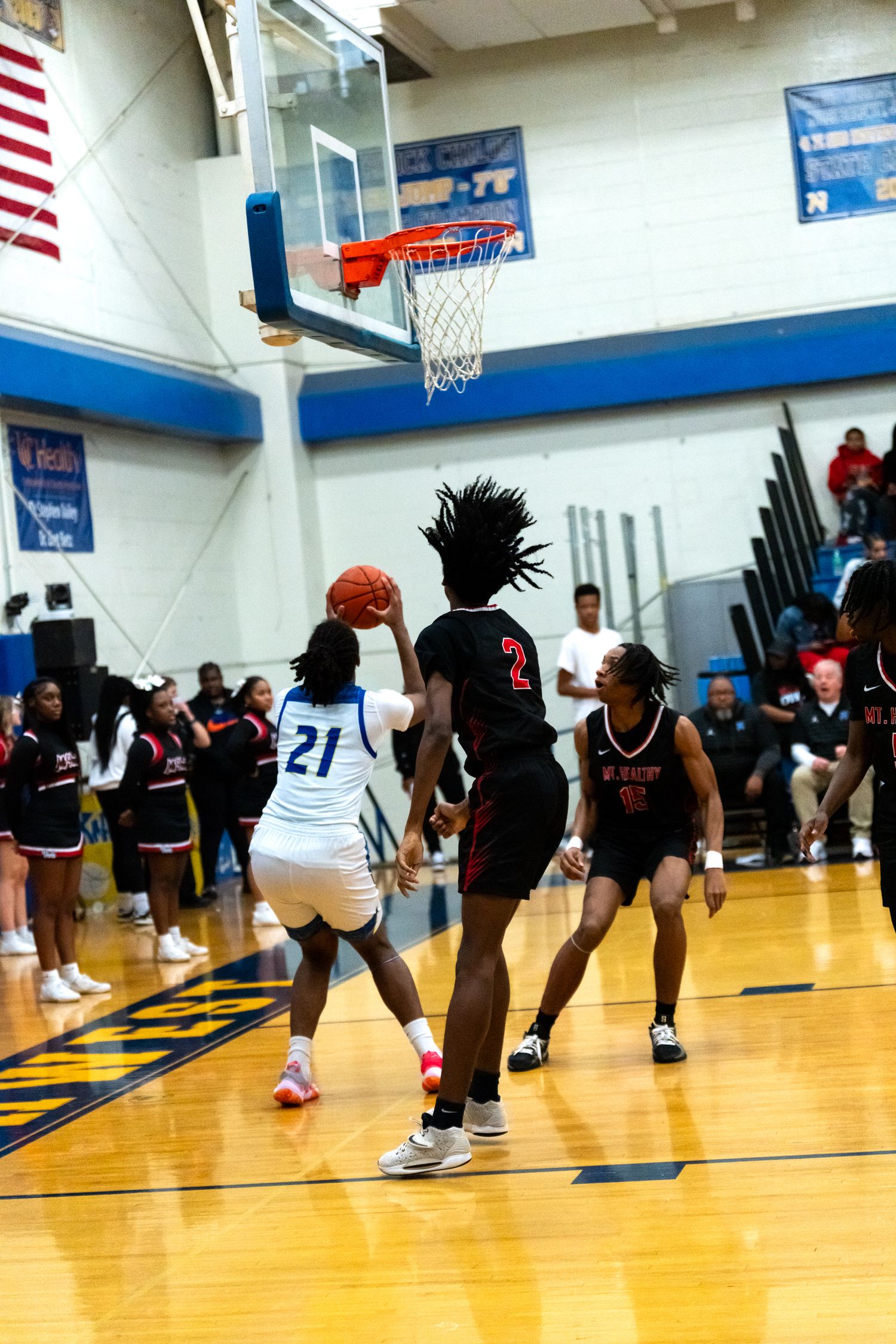 A player in white number 21 jersey attempts a layup against defenders in black during a high school basketball game.
