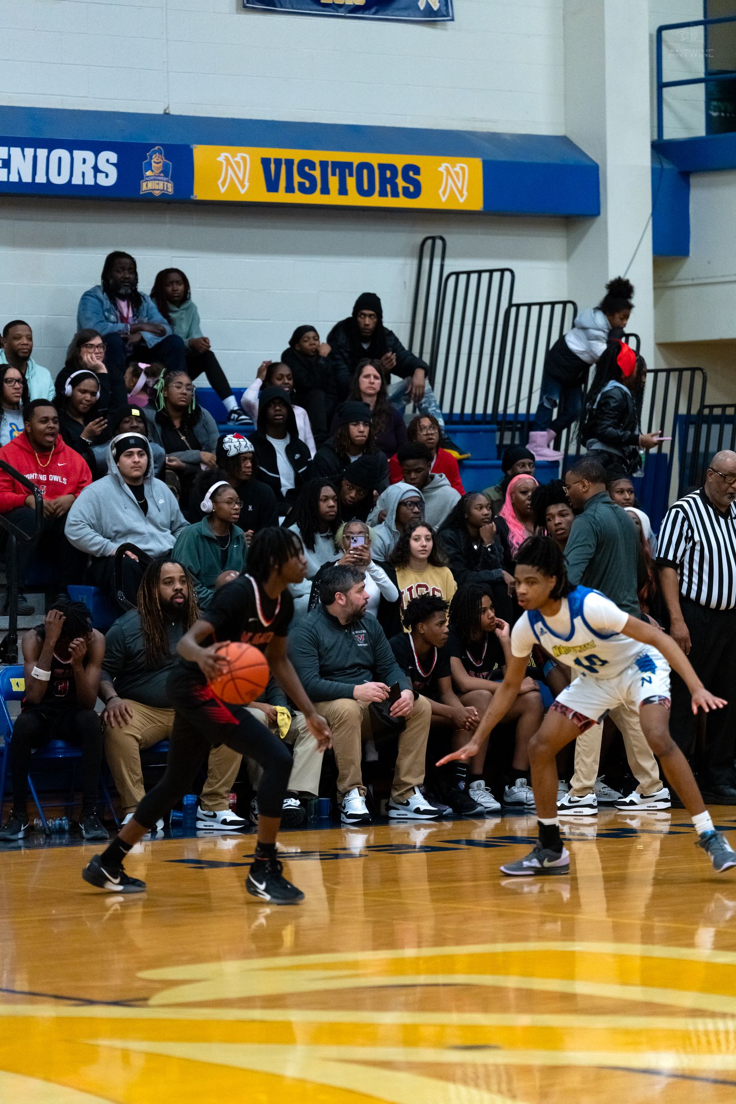 Basketball player in dark uniform dribbles near sideline while defender in white guards during crowded gym game.