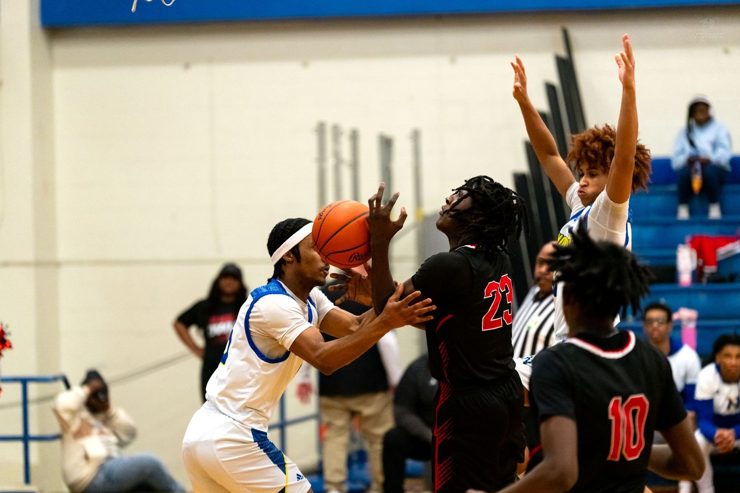 A basketball player in white attempts a shot while defenders in black jerseys contest during an indoor high school game.
