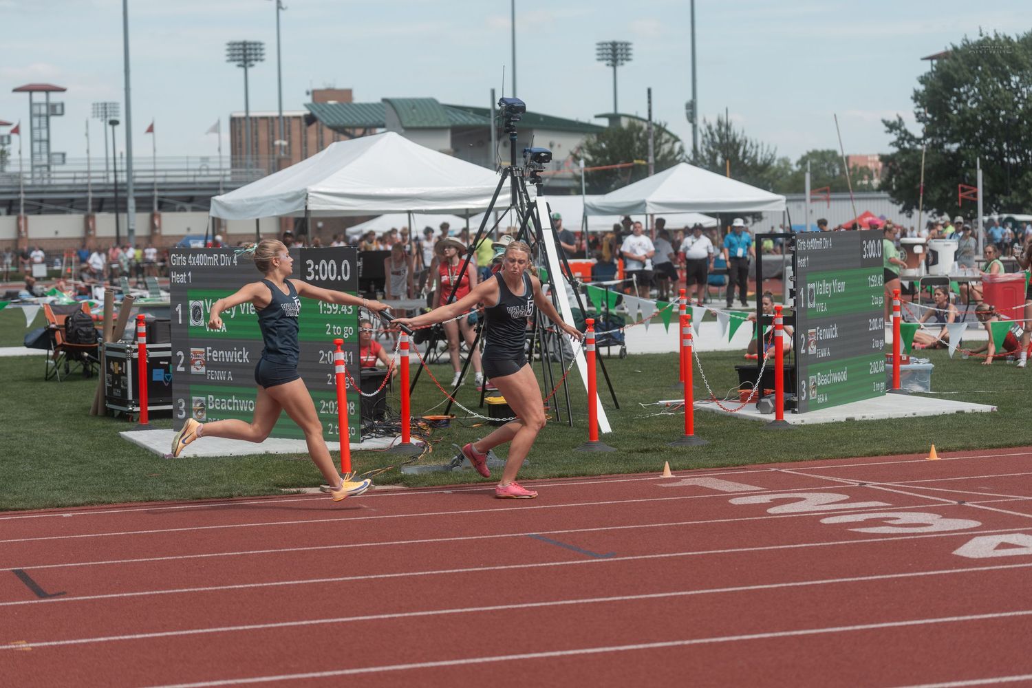 Female relay runners complete a baton handoff during a competitive race at a high school state track meet.