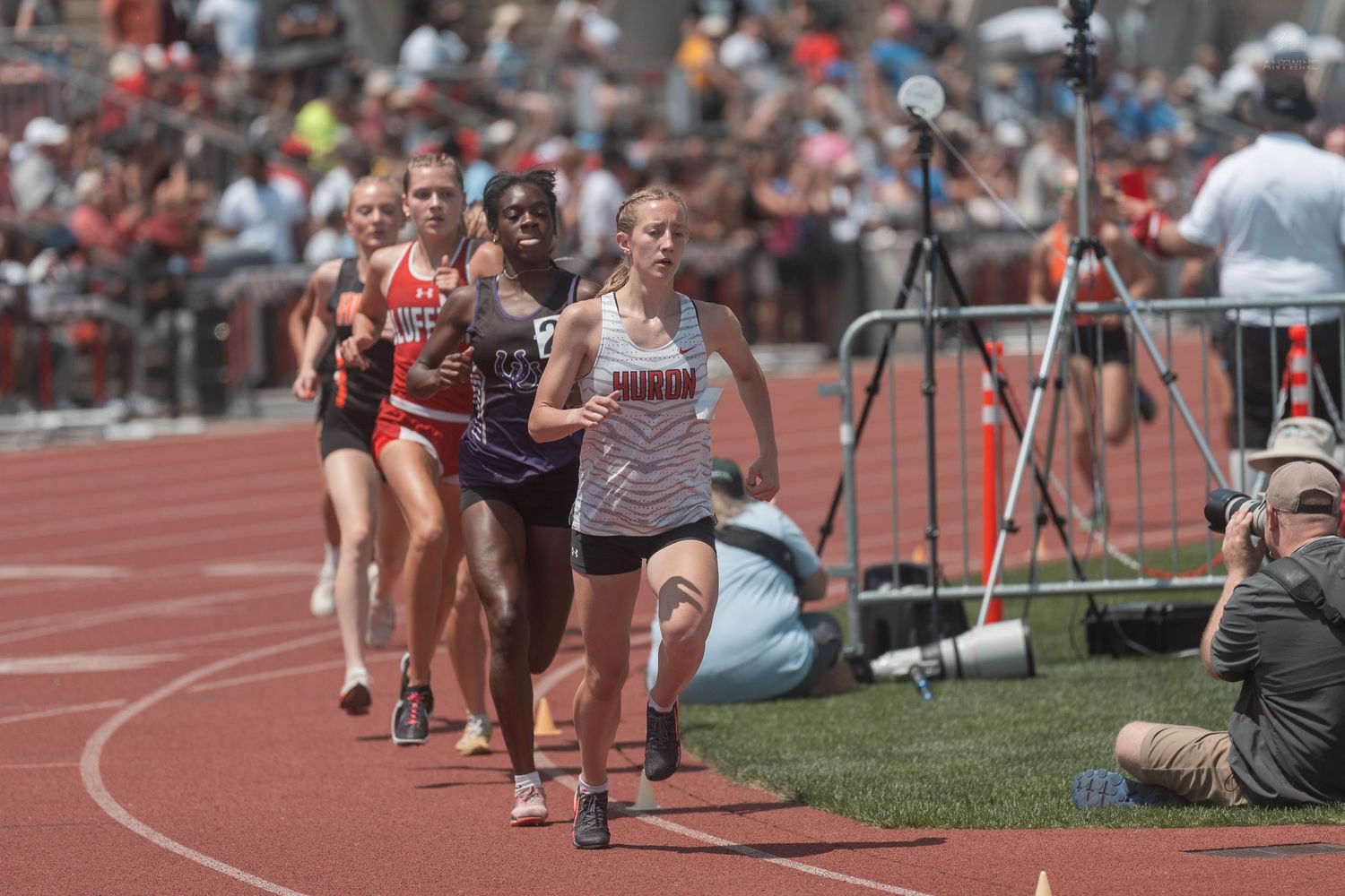 Female athletes race on a red track during a competitive high school track and field meet in bright sunlight.