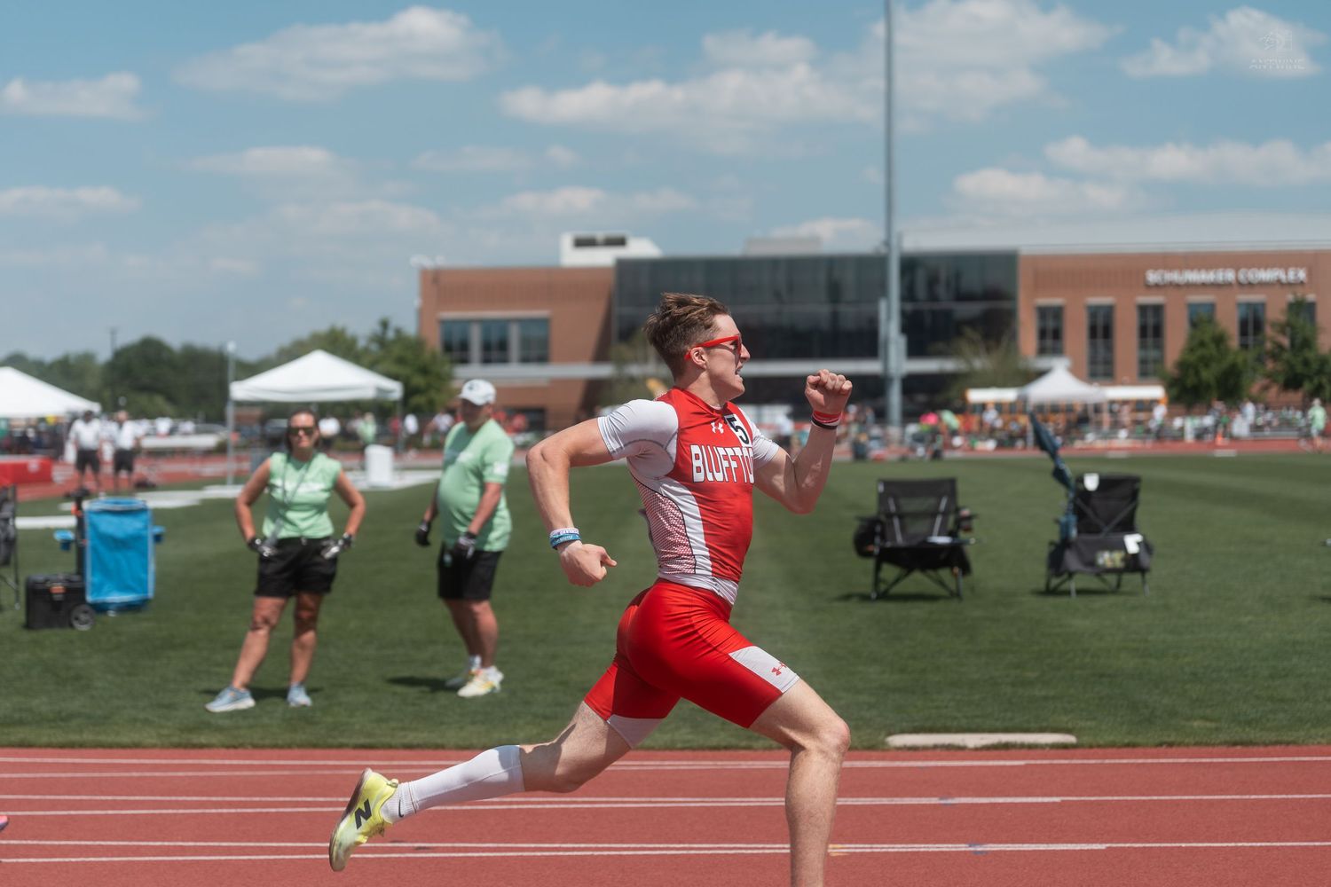 Male athlete in red uniform sprinting on a track during a competitive outdoor track and field meet on a sunny day.